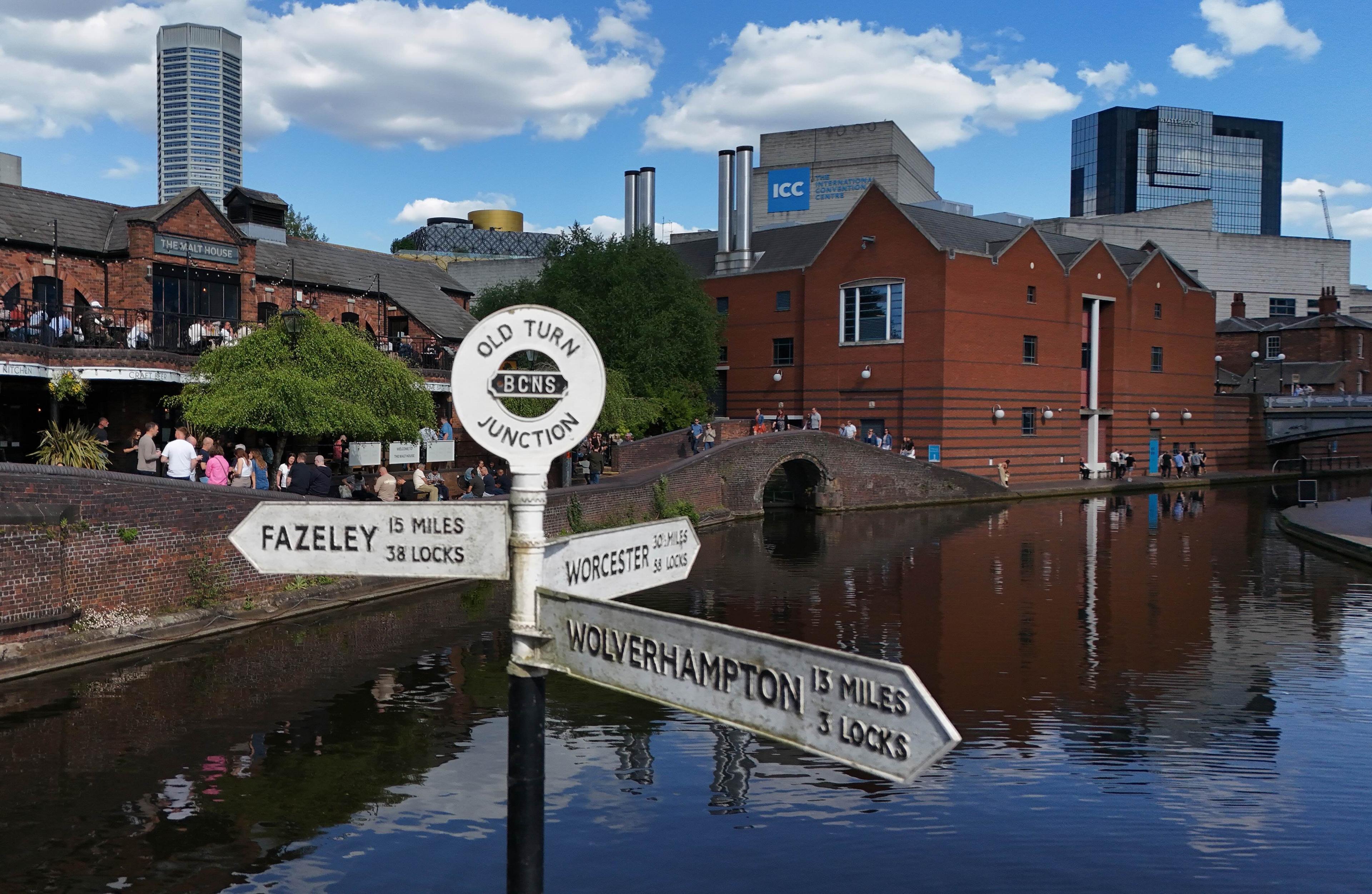 A white canal sign at Old Turn Junction signalling the direction to Fazeley, Wolverhampton and Worcester. Red brick buildings and a bridge can be seen at the canalside