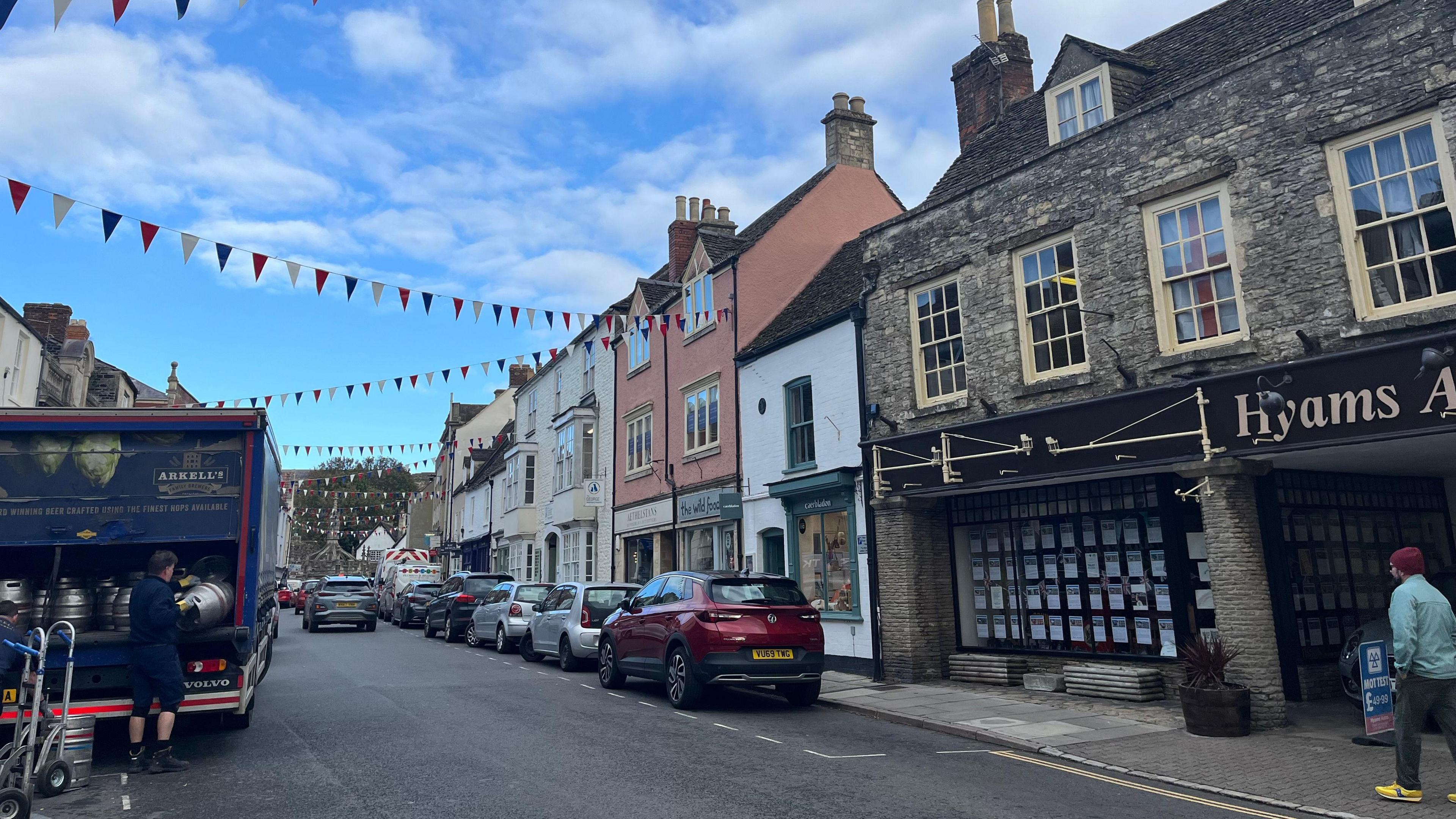 High street with bunting, different colour shop fronts, several cars and a lorry with a man loading vats of alcohol into it. It is a blue sky with some clouds above the street.