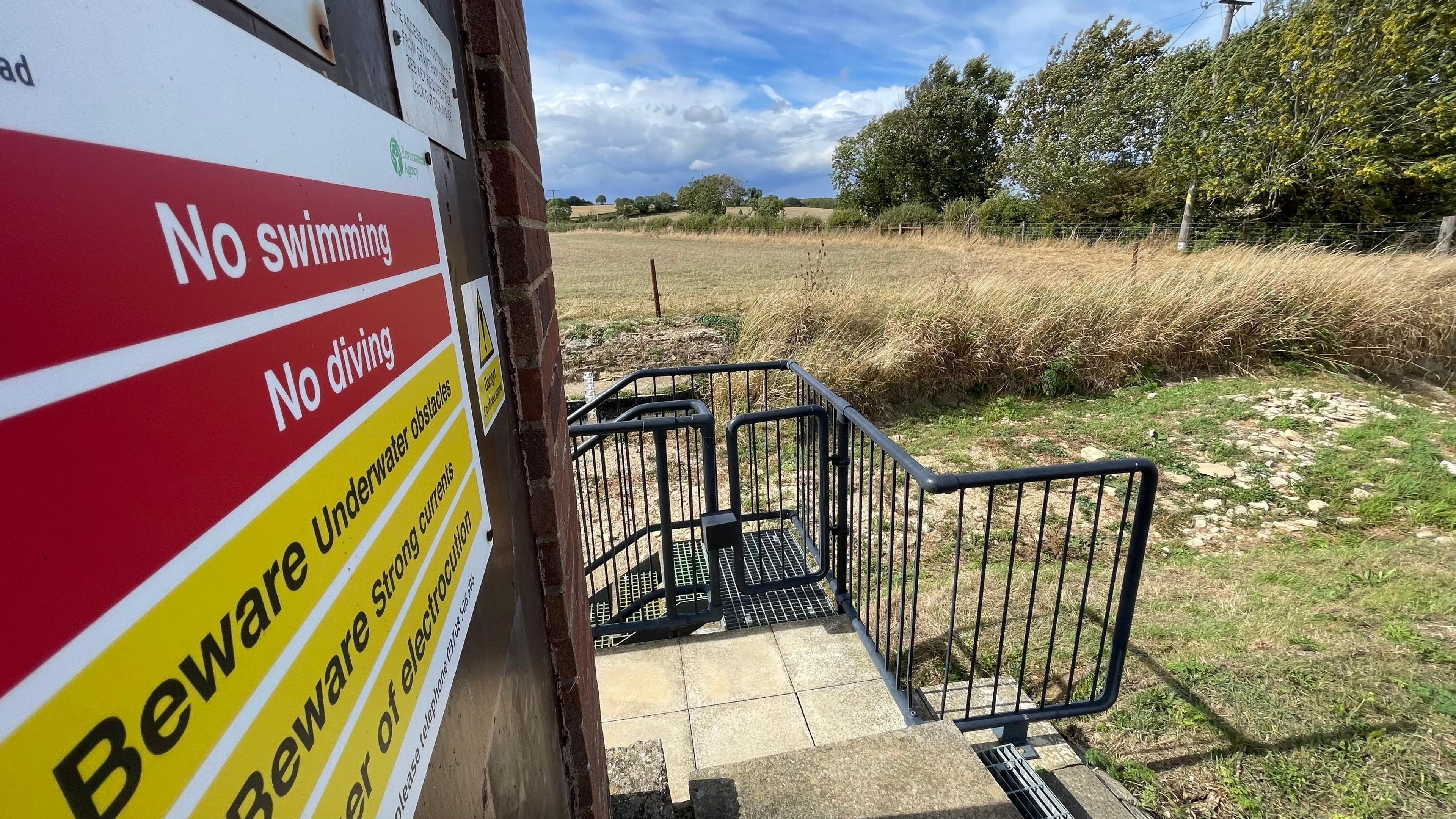 Signs in the foreground warn of 'no swimming' and 'no diving' while in the background there is a platform leading down to a dried-up river bed strewn with boulders.