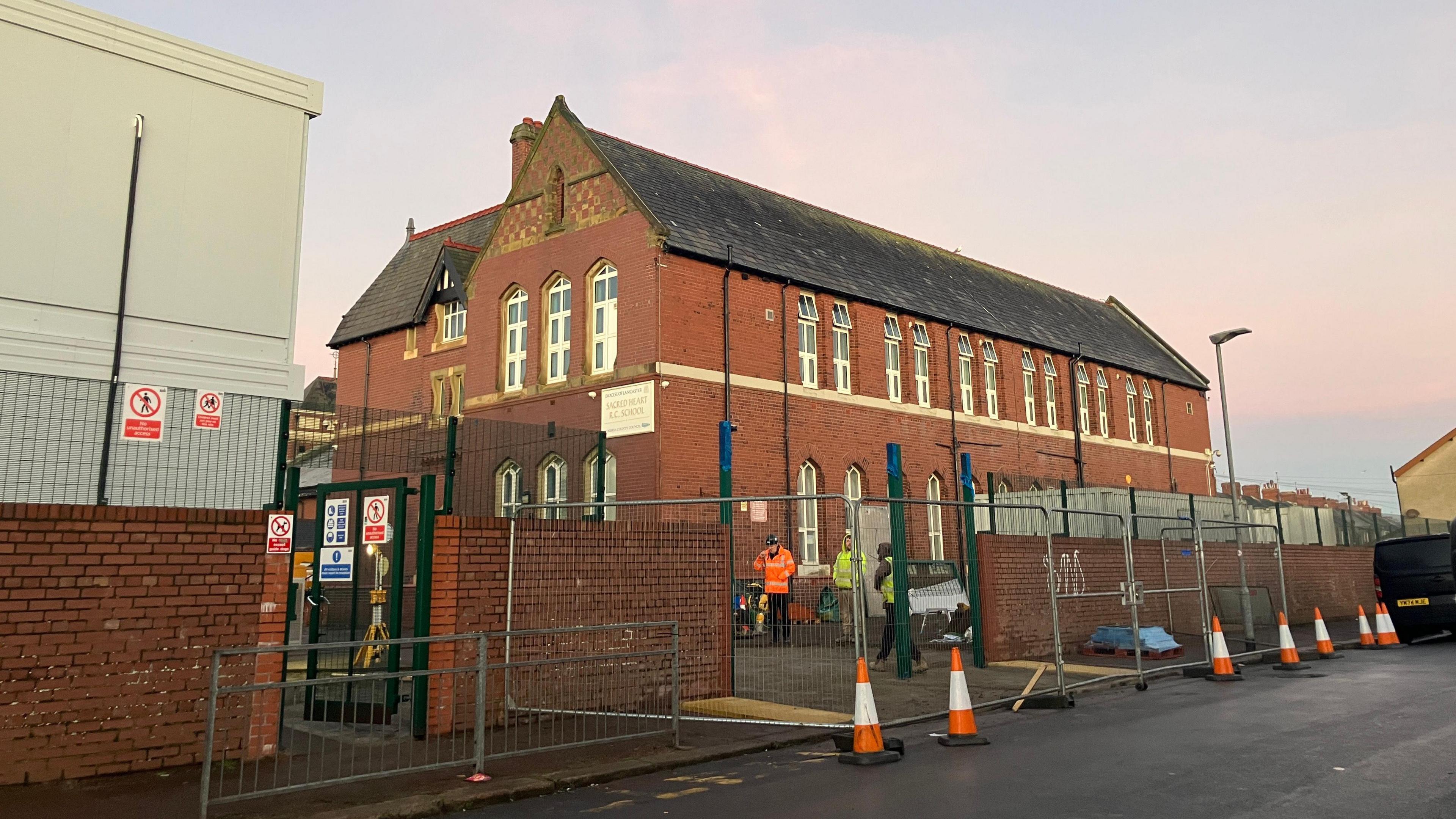 Sacred Heart Catholic Primary School is a red-brick school building. There are two-storeys of windows. Workers in high-vis jackets are walking around the exterior. Temporary metal fences have also been erected.