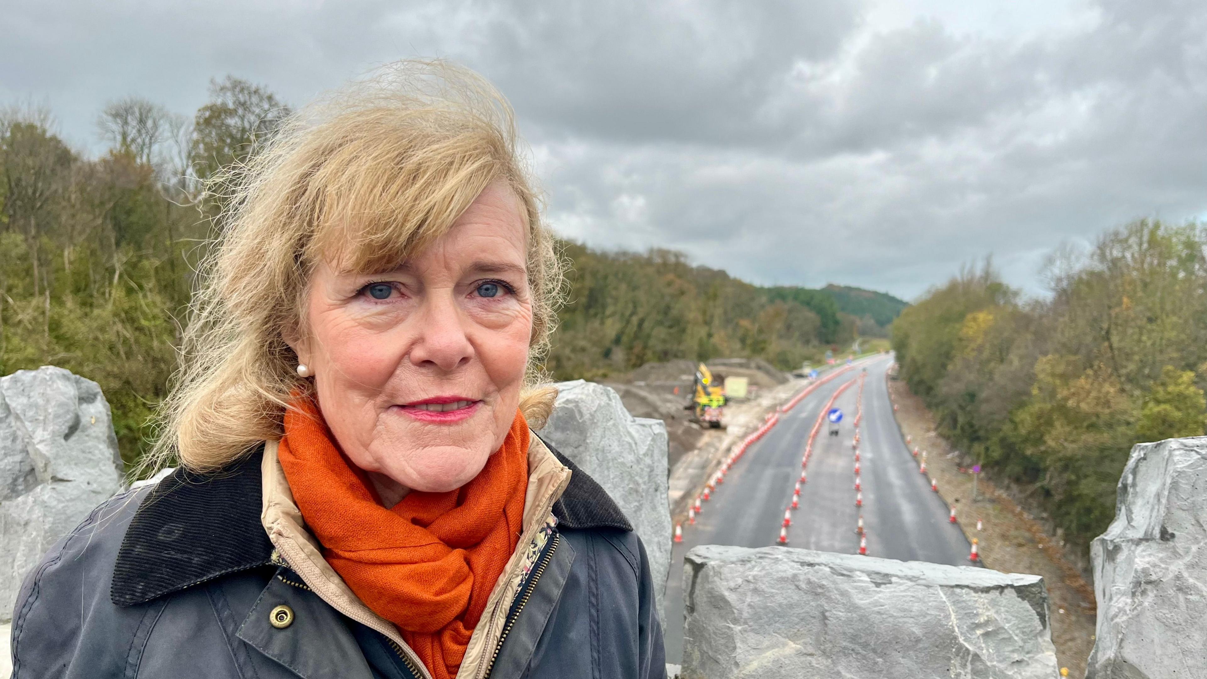 Andrea Davis standing on the West Buckland bridge with the North Devon Link Road behind - she is wearing an orange scarf and a grey jacket with corduroy collar.