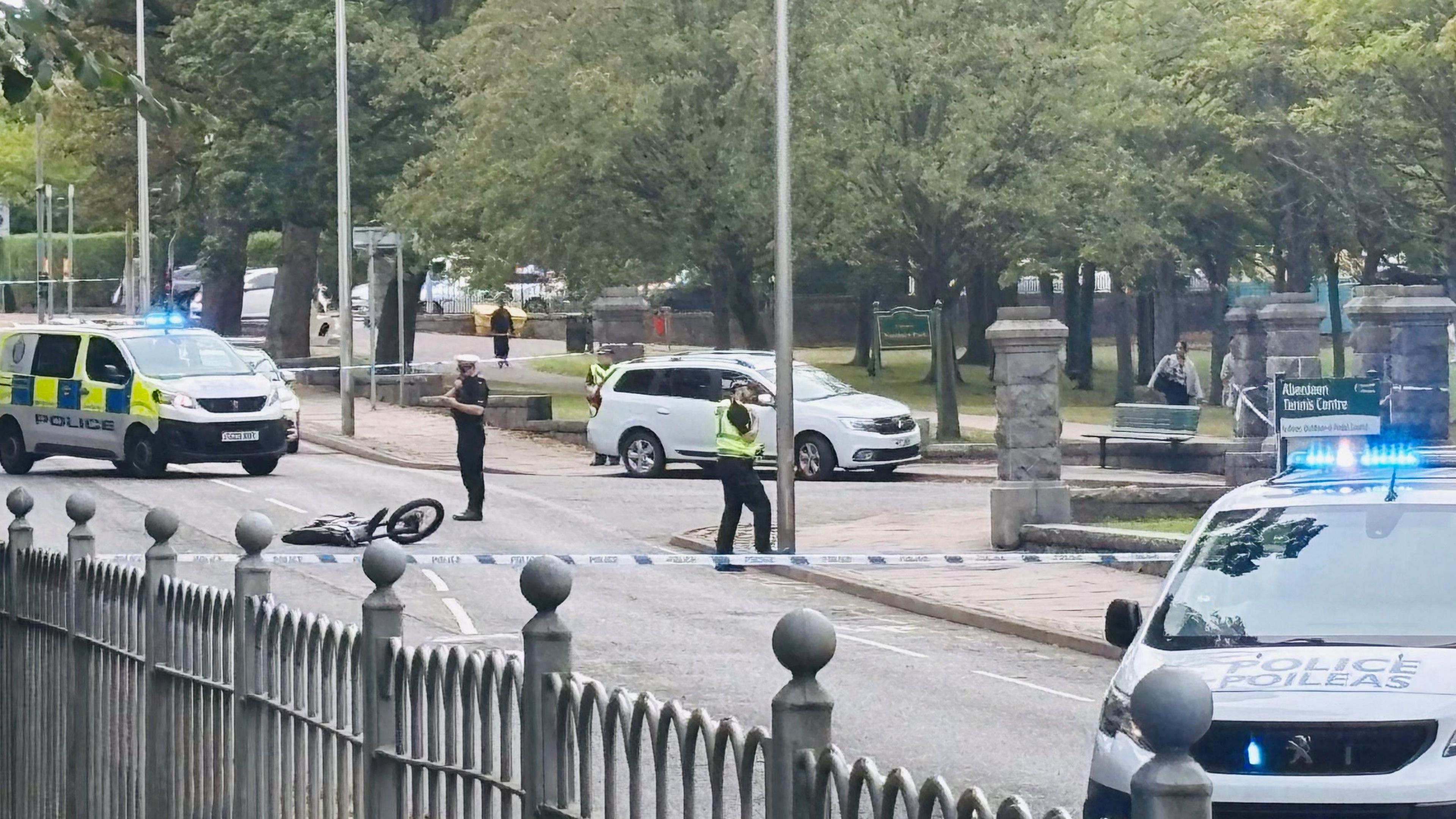 Police vehicles, police tape and police officers at scene of crash bike on road, in between two parks.