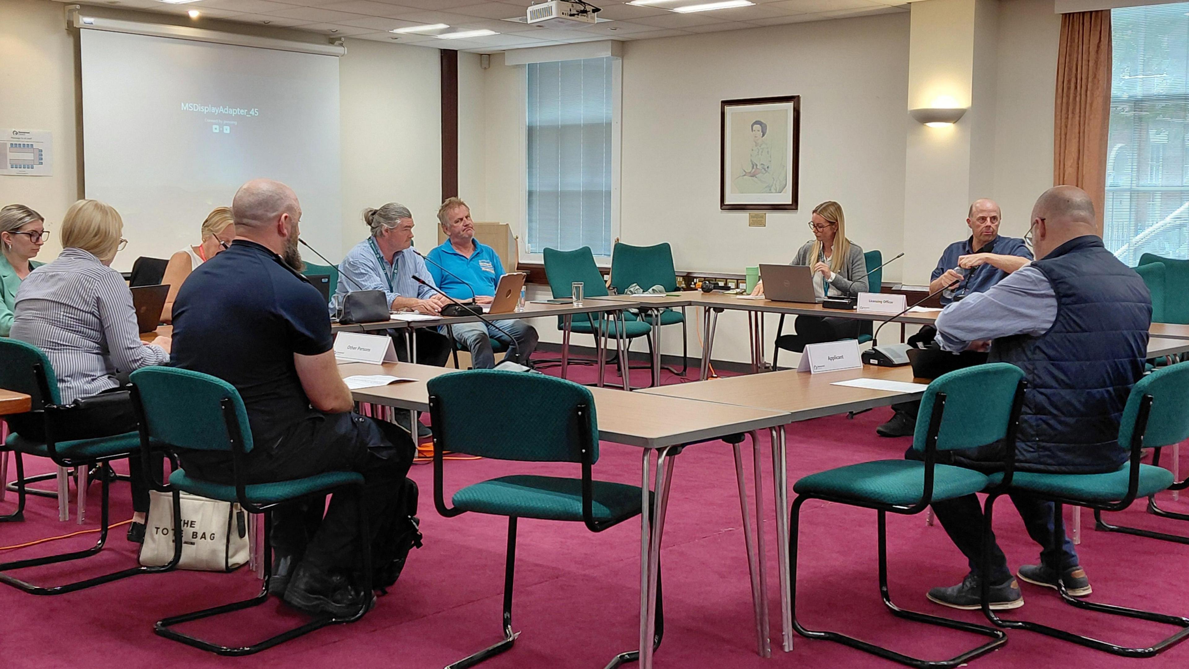 People seated around a table arrangement in a conference room with red carpet and green chairs. A projector screen at the front displays ‘HDMI Searching…’ as participants engage with laptops and documents. Nameplates and framed wall art suggest a formal meeting or workshop setting.