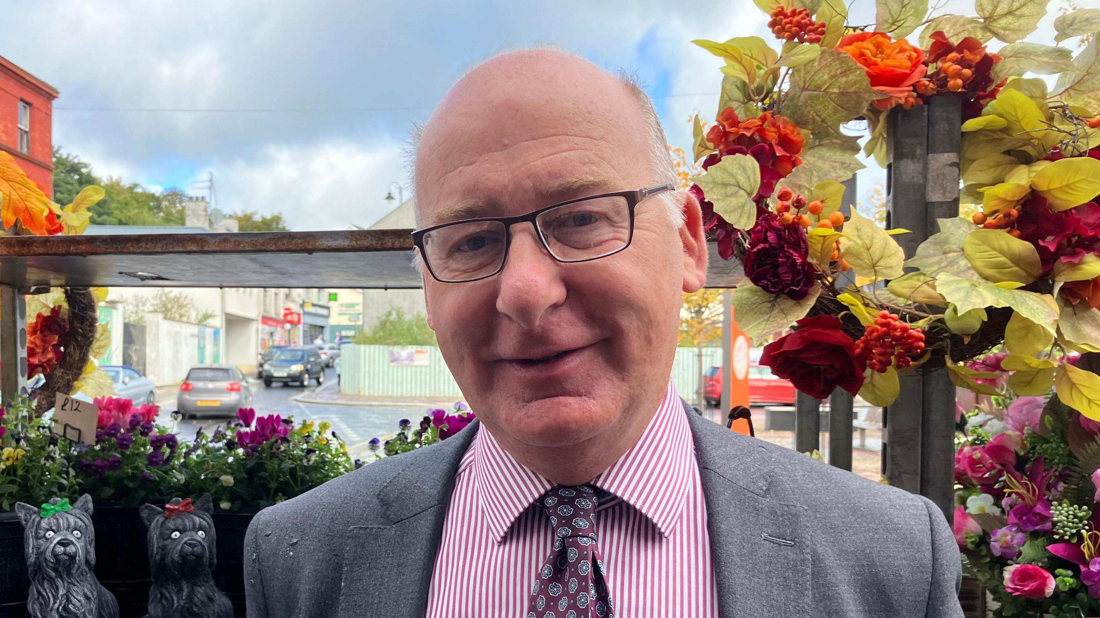 A man in a grey suit jacket looks into the camera. He is wearing a pink striped shirt, a purple patterned tie and dark rimmed glasses. There are wreathes in red, green and orange hanging behind him and two black statues of dogs. In the distance you can see cars travelling on a small road. 