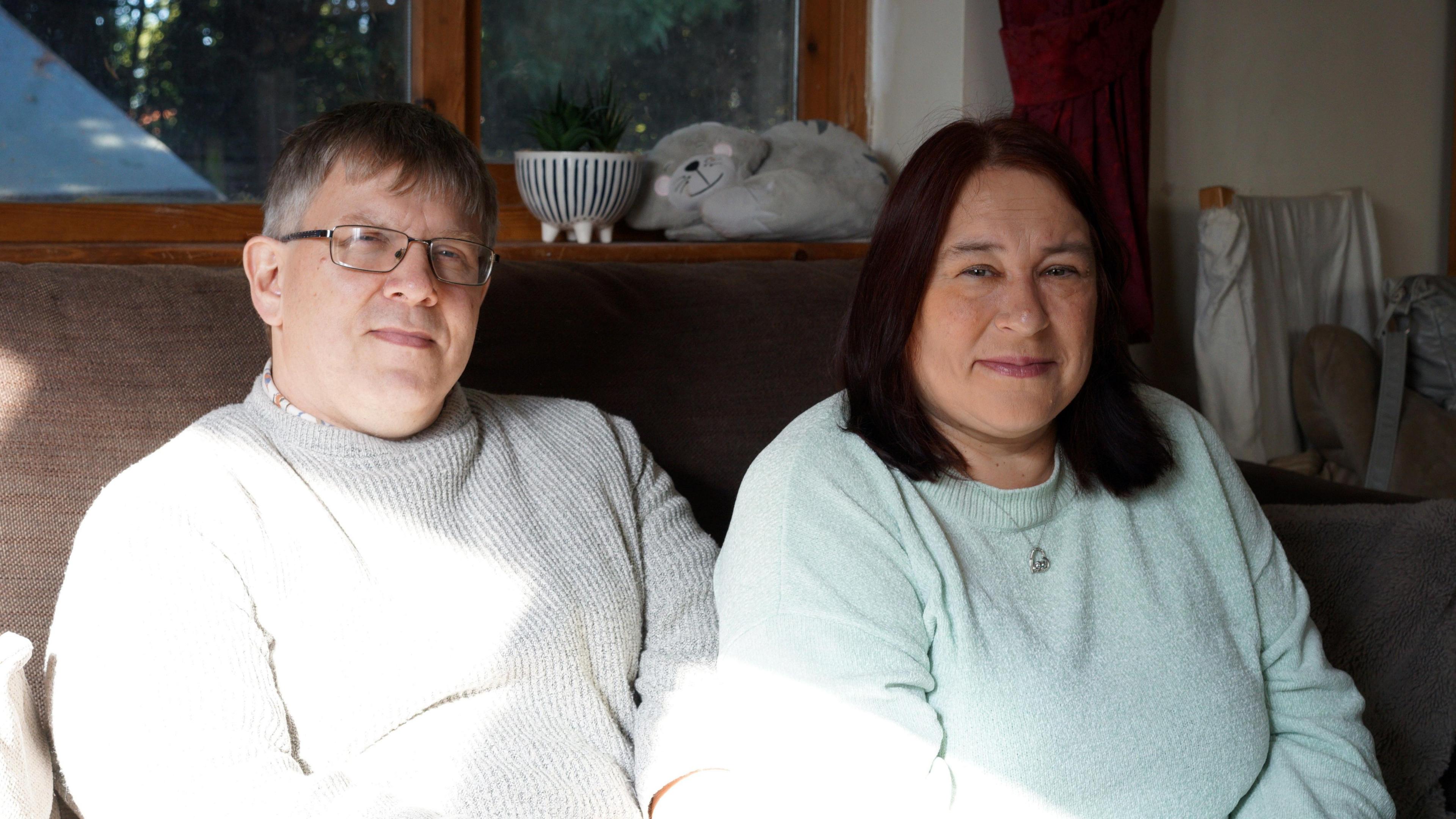 Adrian and Judith Winckles are sitting on a brown sofa in their living room, looking at the camera, smiling faintly.