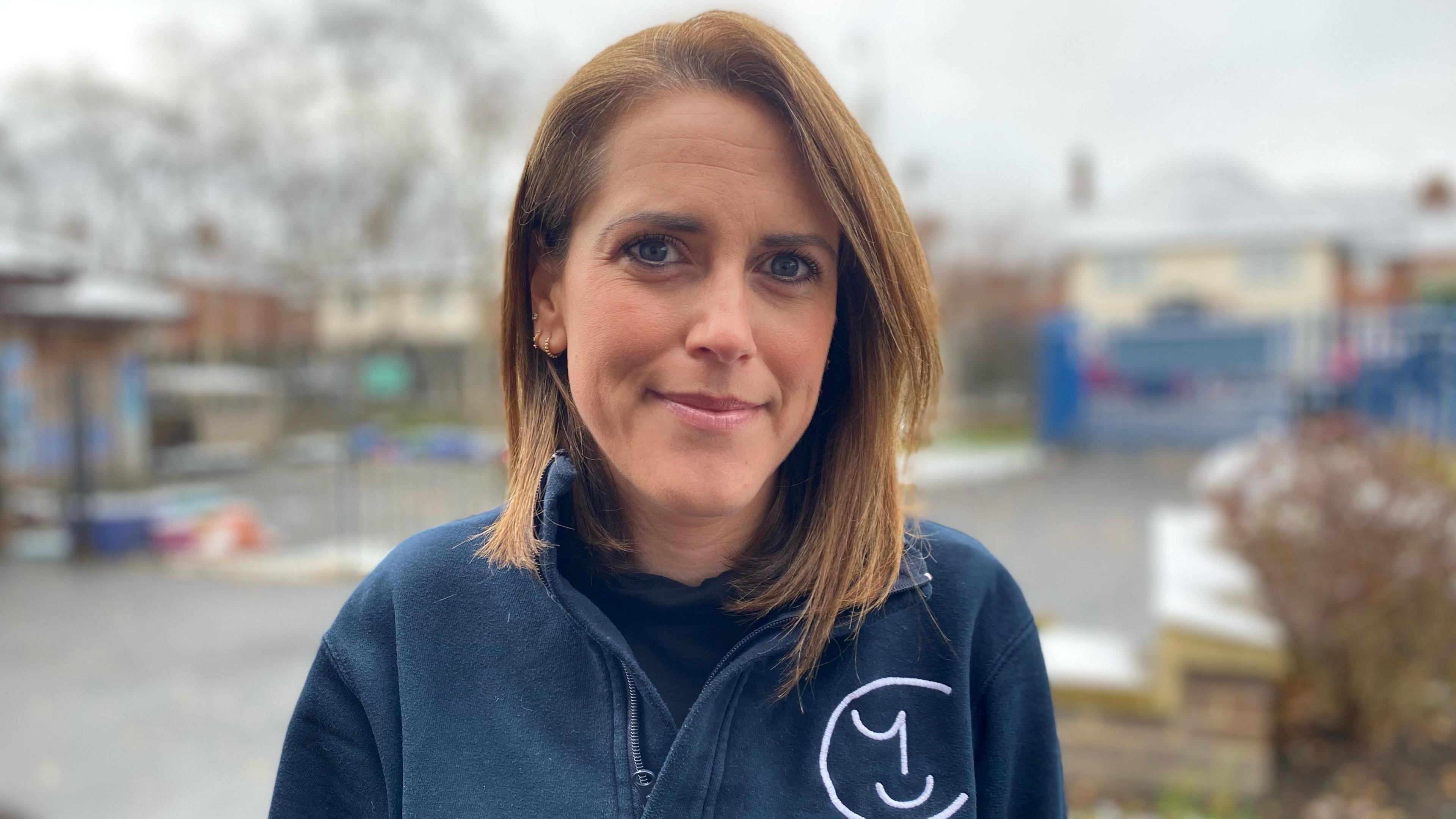 Ms Farrington has shoulder length copper hair and wears a navy zip hoodie with the charity's logo. The hair on her left is tucked behind her ear, making three gold earrings visible on her ear.  