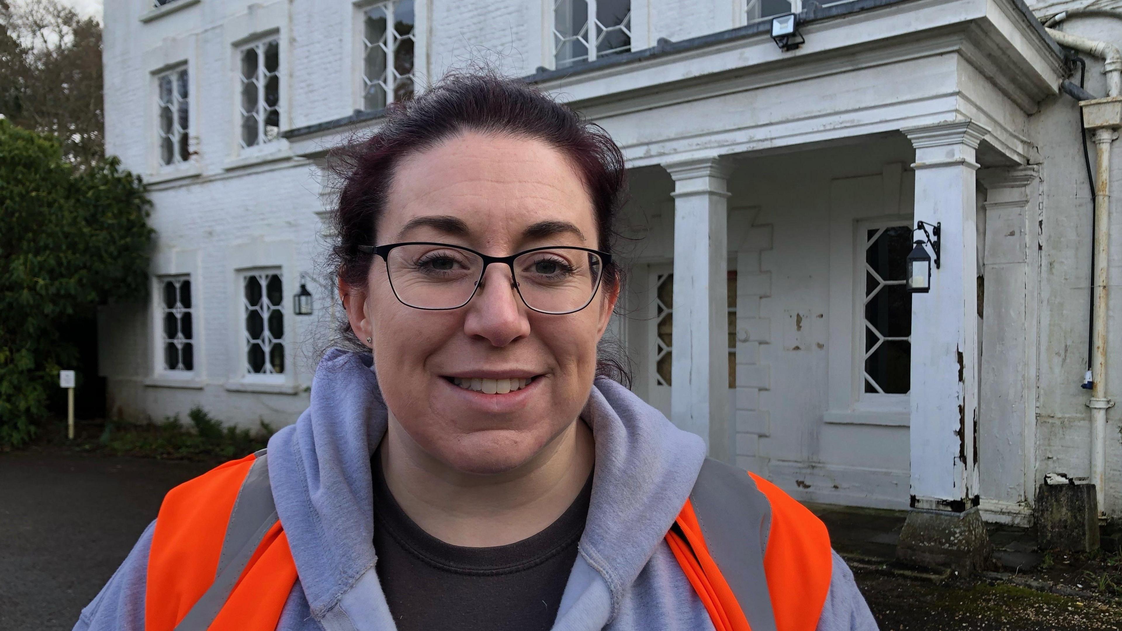 Emma Stevens stands outside an old building with peeling white paint. She is wearing a grey hoodie, glasses and a fluorescent orange jacket.