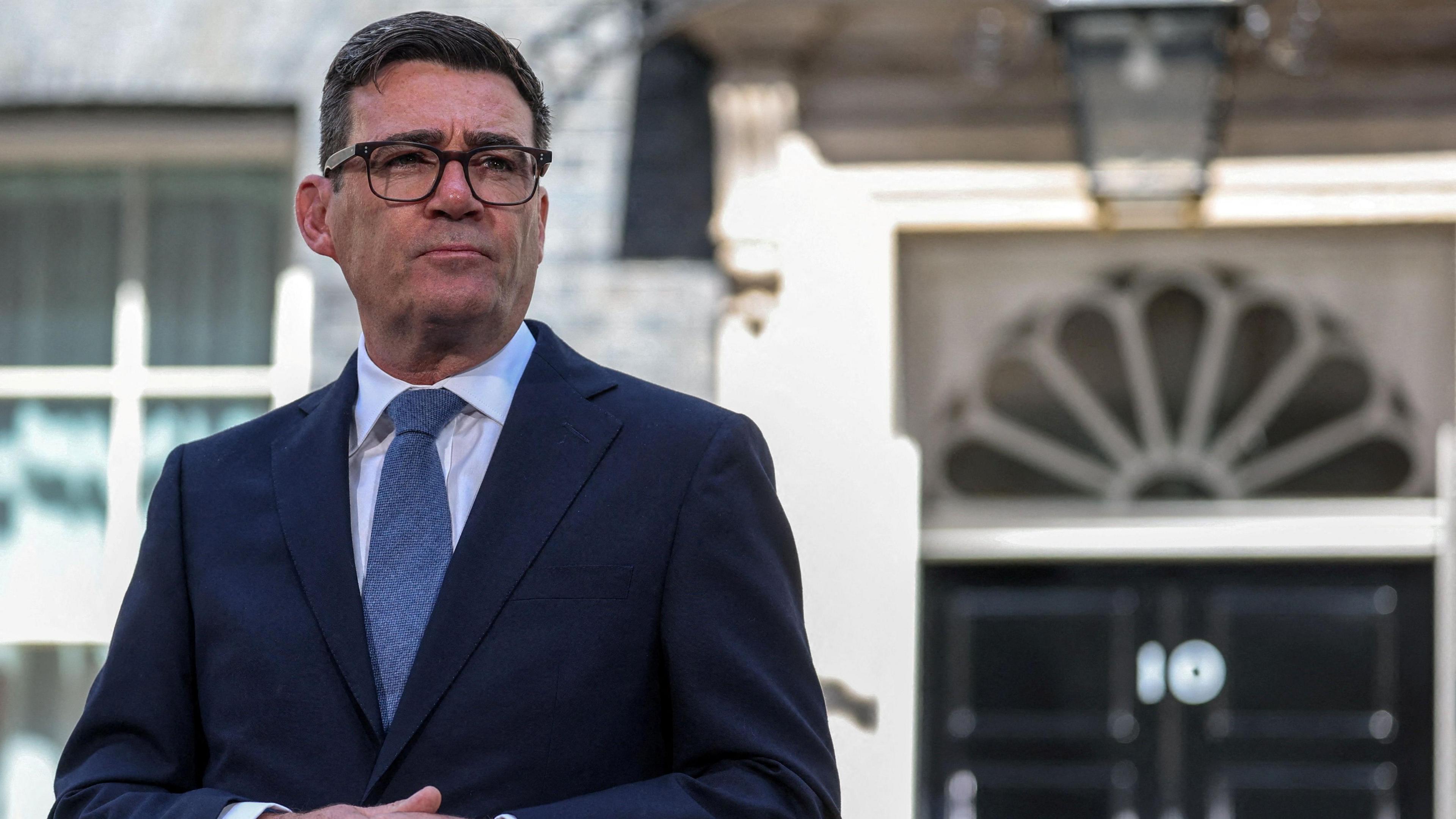 Andy Burnham standing outside No 10 Downing Street. He is wearing glasses, a white shirt, blue tie and navy suit. 