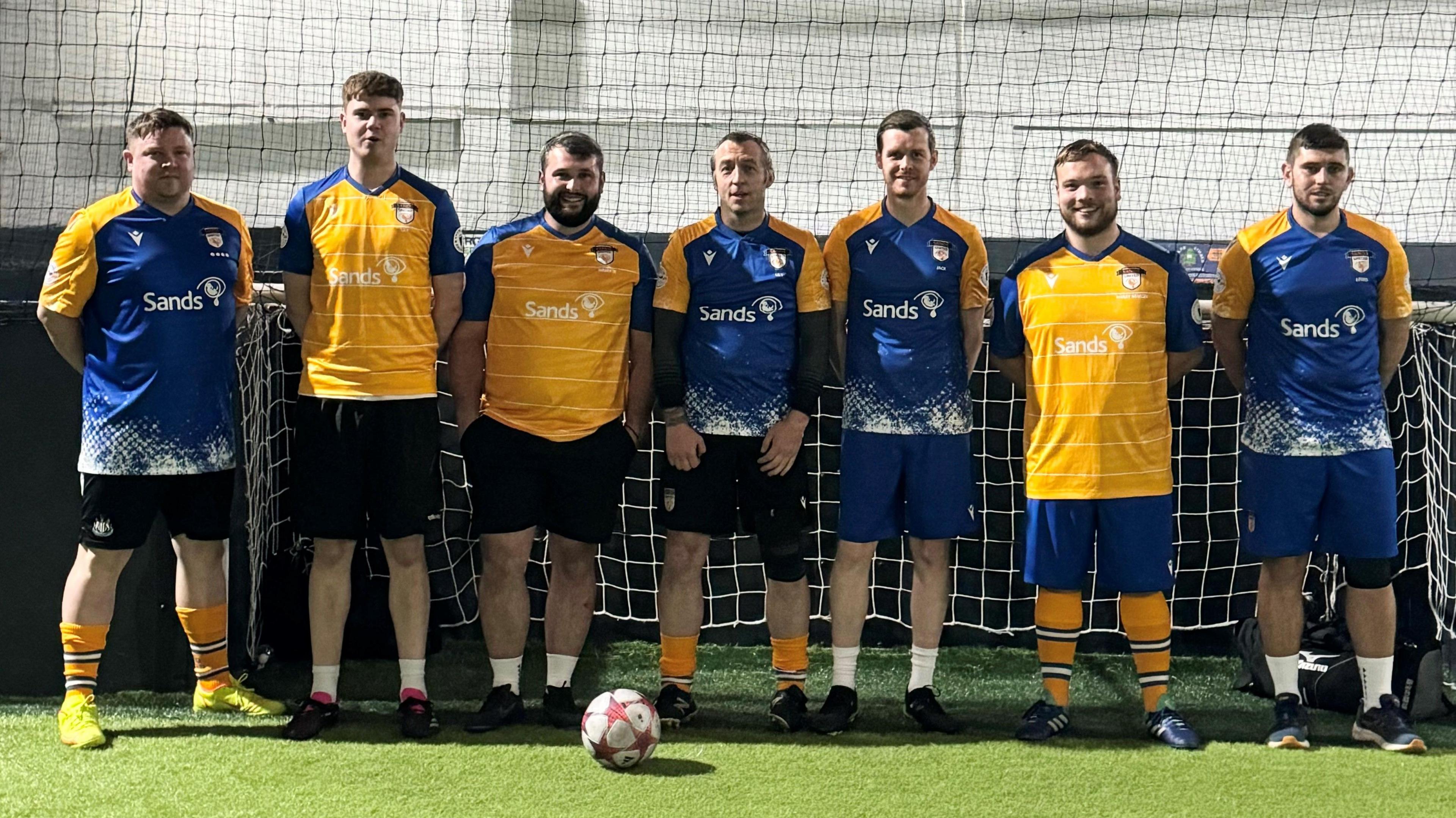 A football team lines up for a photo in front of a football net. There are seven men, wearing blue and yellow kit. They are stood on green grass and a football is in front of them.