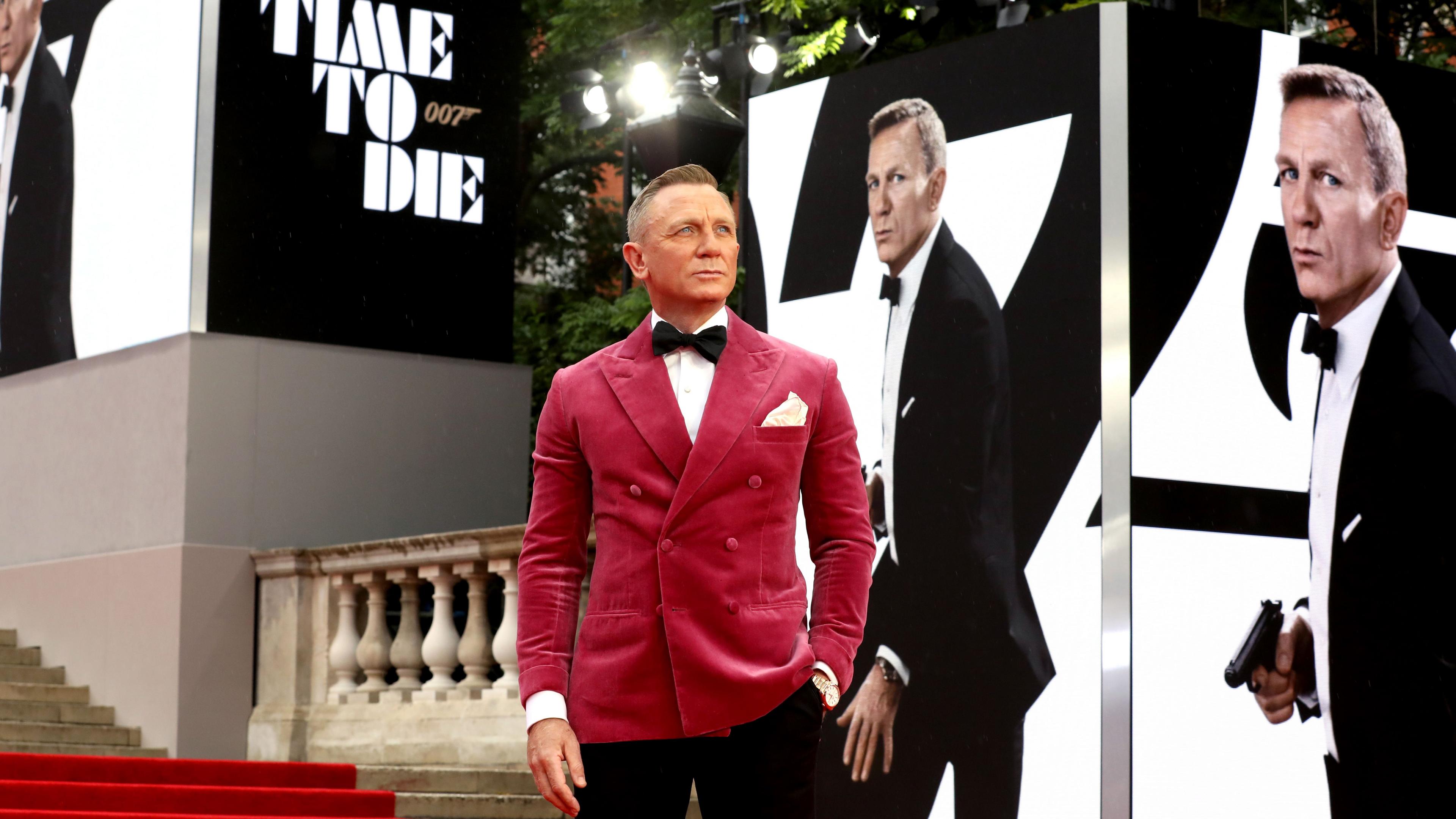 Daniel Craig appears at the premiere of No Time to Die (2021) wearing a striking berry suit jacket and posing in front of pictures of himself playing James Bond