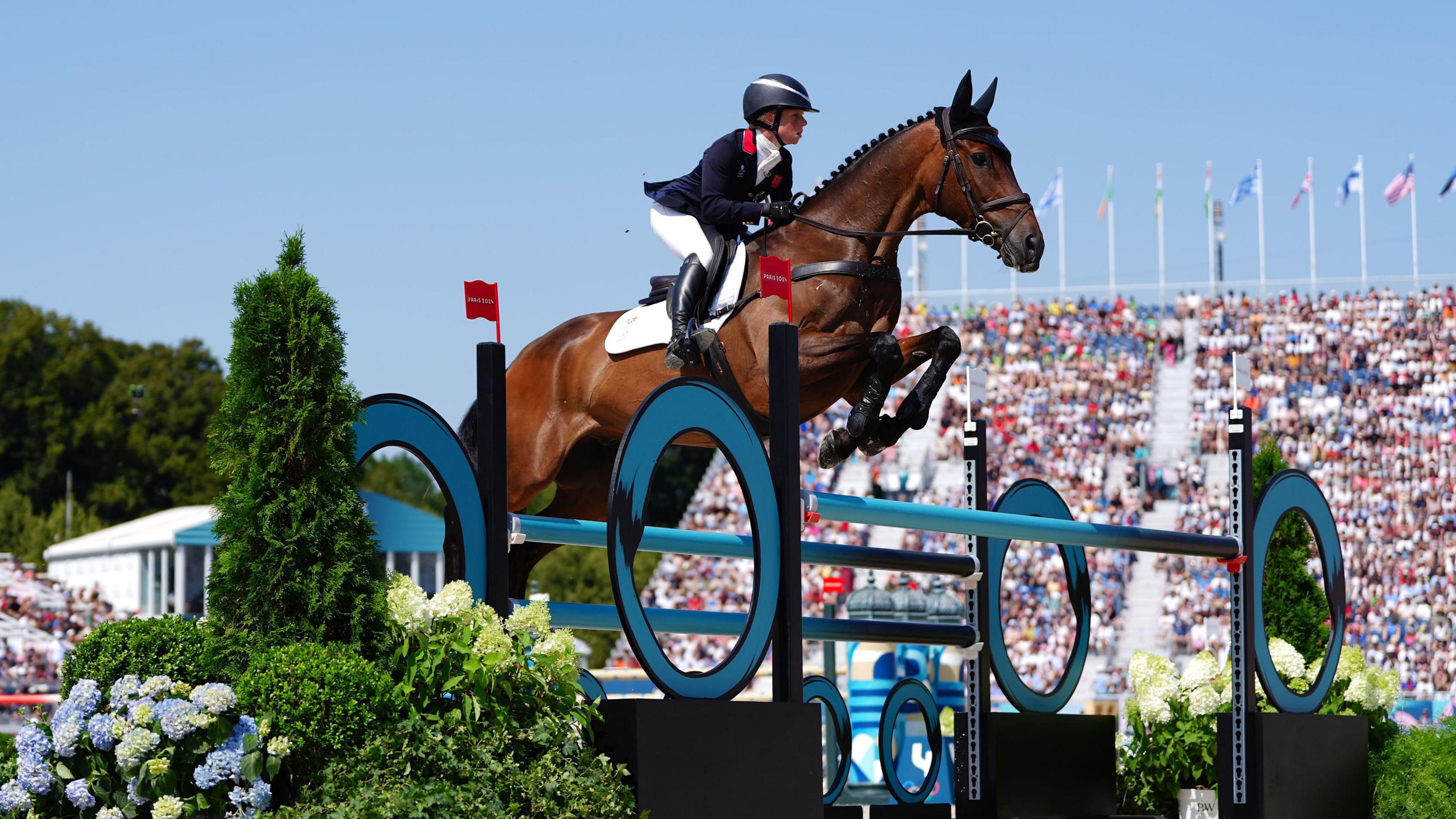 Ros Canter rides her horse over a large jump in the arena, there are blue skies and a large crowd of spectators watching.