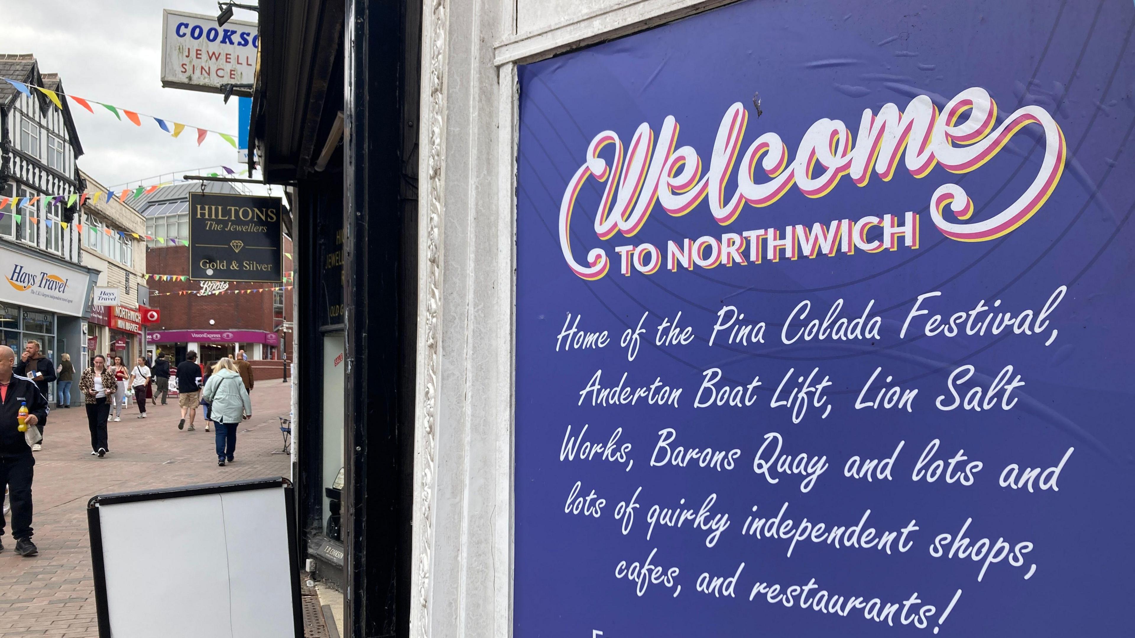 A large purple-and-white sign in a high street doorway which reads: "Welcome to Northwich, Home of the Pina Colada Festival, Anderton Boat Lift, Lion Salt Works, Barons Quay and lots of lots of quirky independent shops, cafes, and restaurants!"