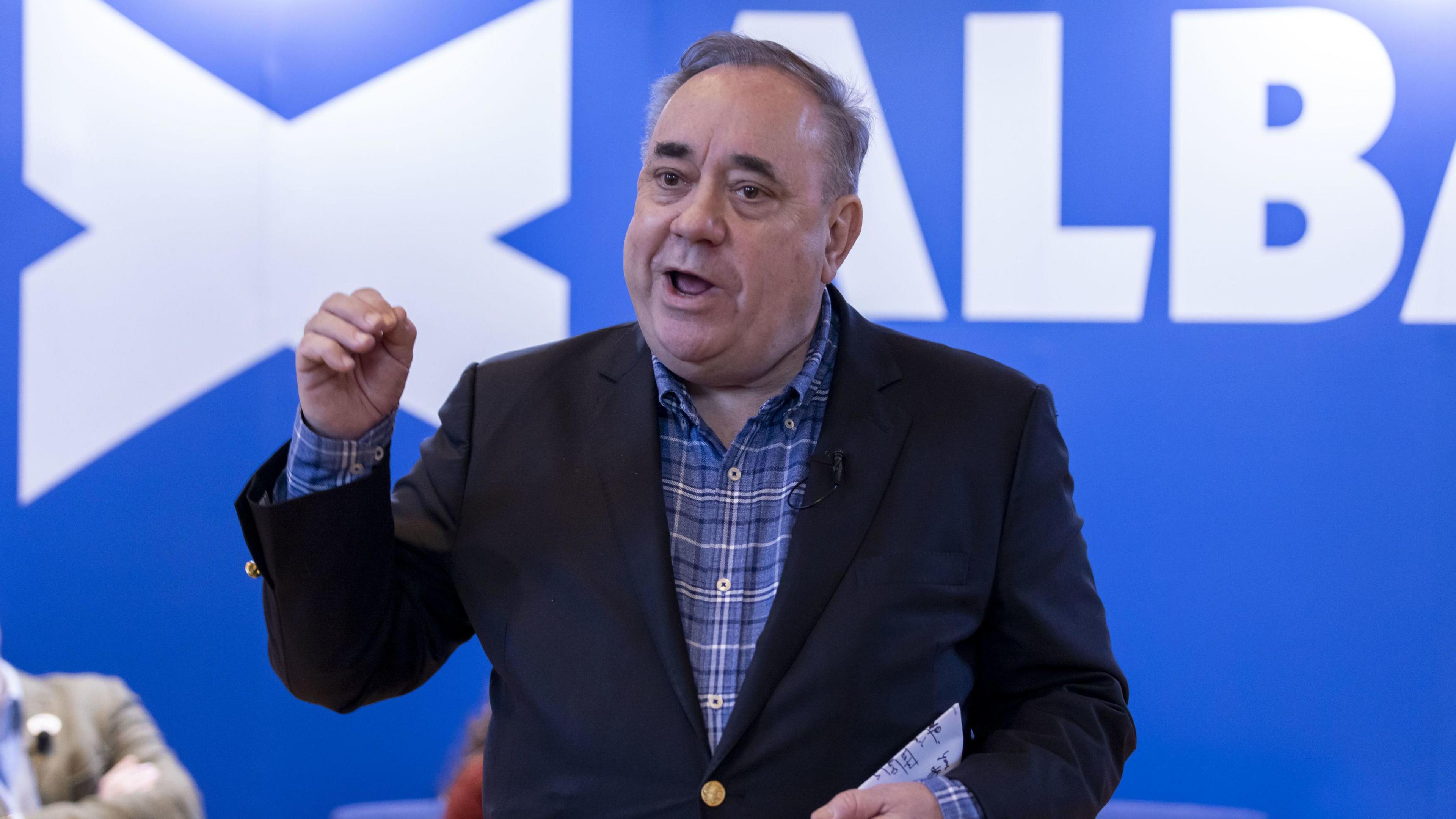 Alex Salmond, wearing a blue, white and dark checked shirt, delivering a speech in front of a blue background featuring the Alba logo in white.