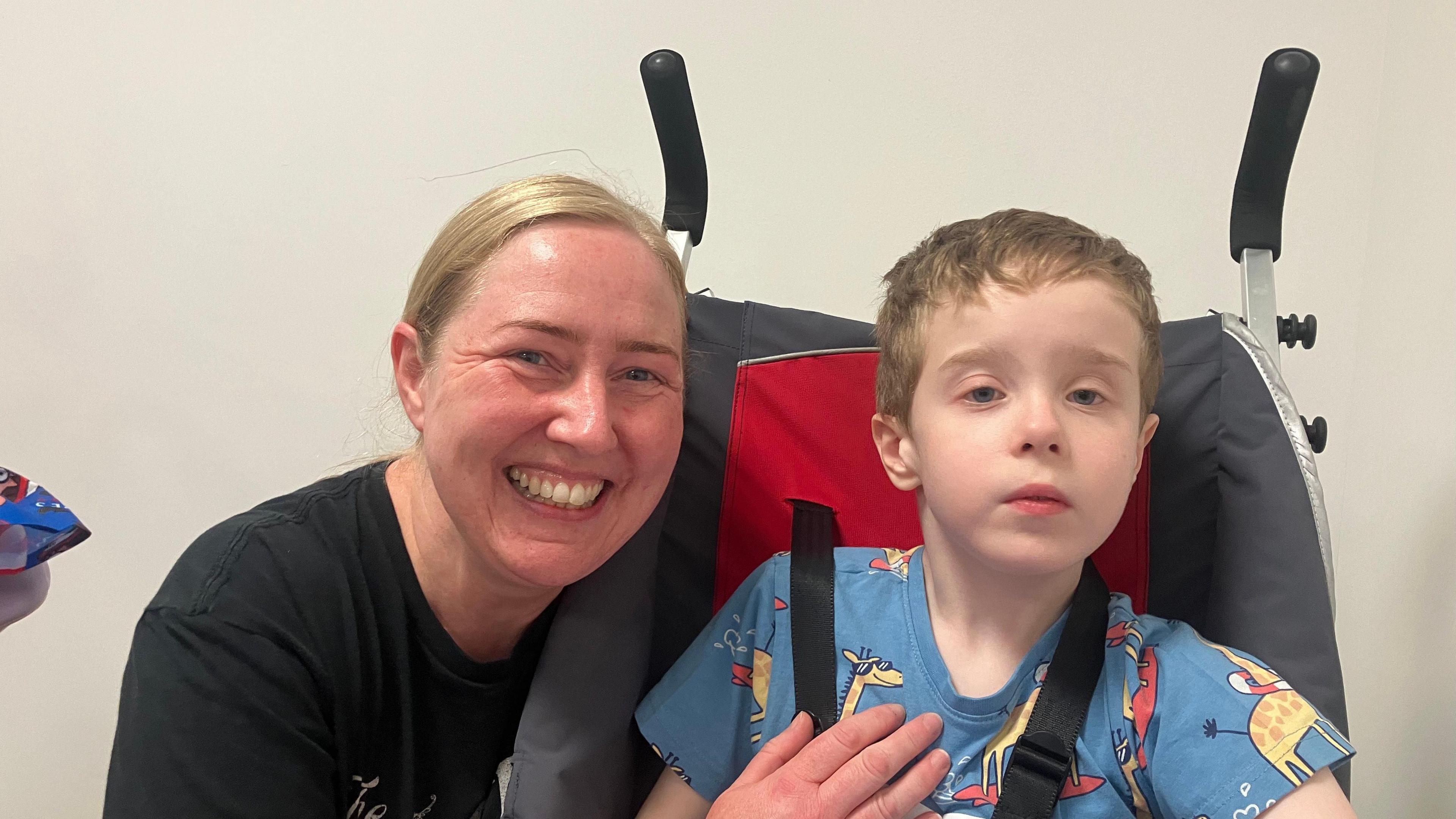 A woman with long blonde hair kneels down and smiles at the camera, alongside her son, who has short blonde hair and sits in a wheelchair, wearing a blue t-shirt. There is a white wall behind the pair. The woman, who wears a black t-shirt, is on the left. 