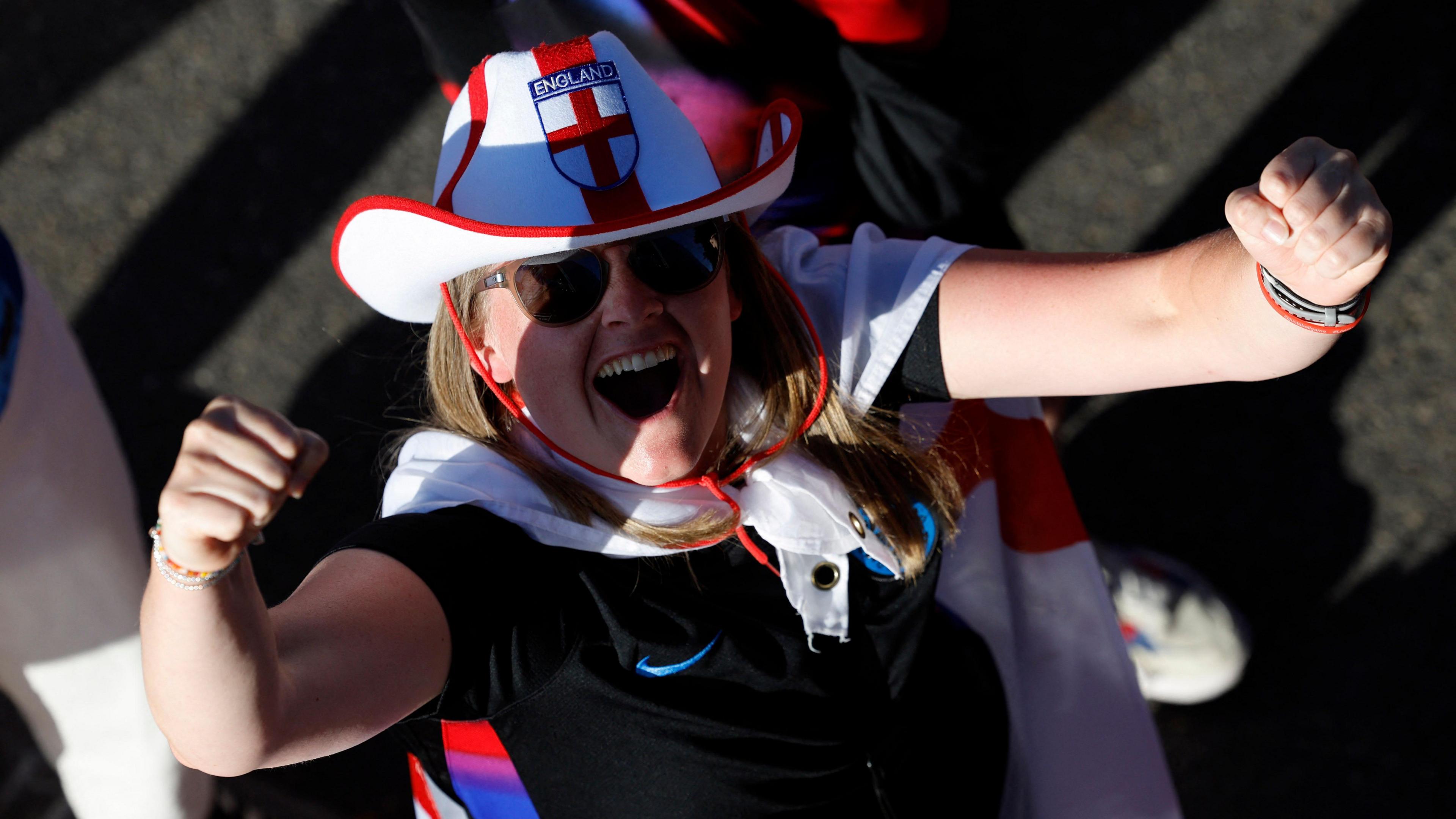 An England fan wearing sunglasses, a red and white England cowboy hat with a flag tied around their neck looks up into the camera punching their arms in the air in celebration.