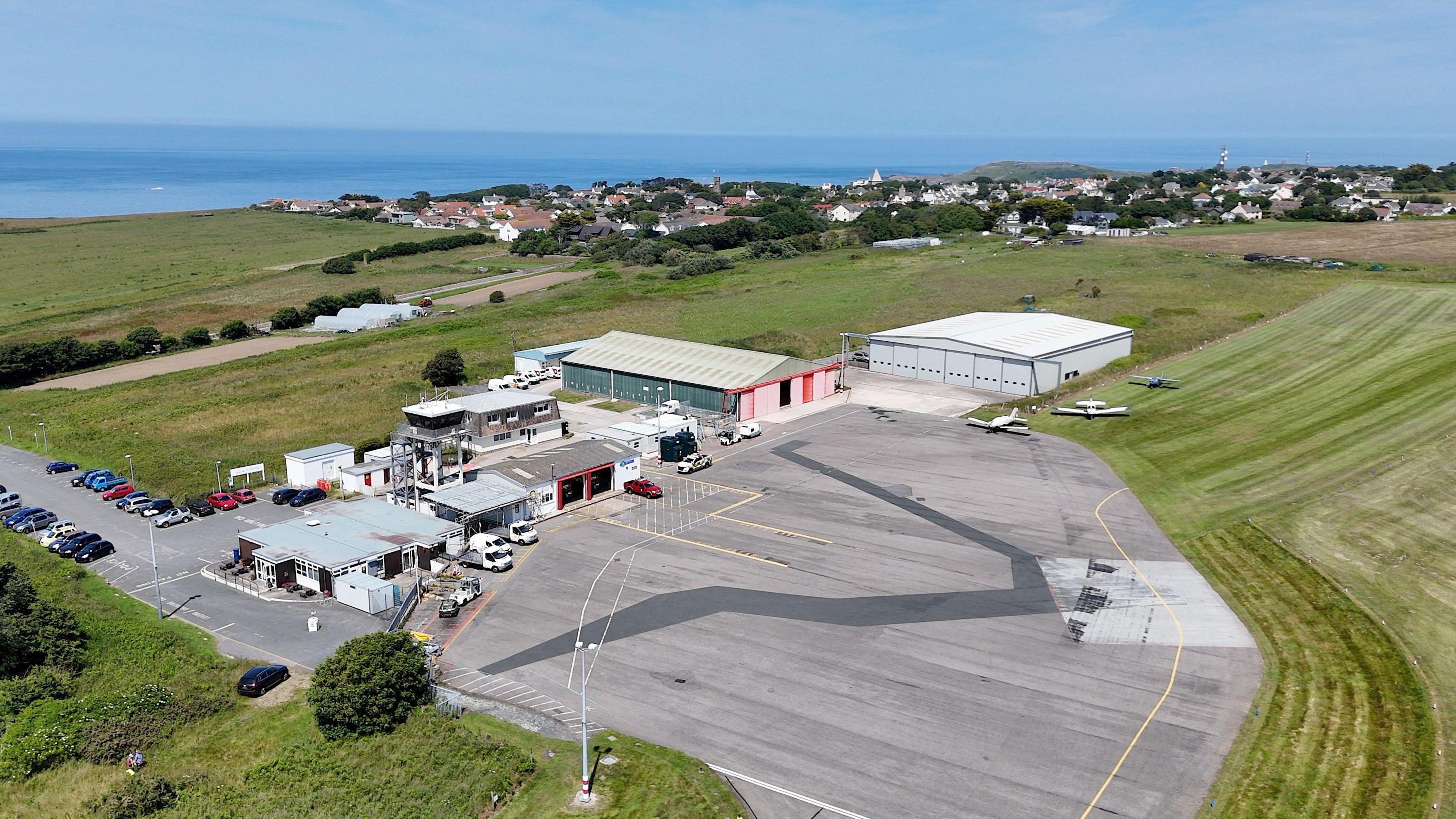 Aerial view of Alderney Airport. Five small buildings. There is a car park being them buildings. The sky is blue. The sea is a darker blue in the background. There are three planes next to a building on the right. There is are fields around. There is a tarmac in front of the building in the middle of the green fields.
