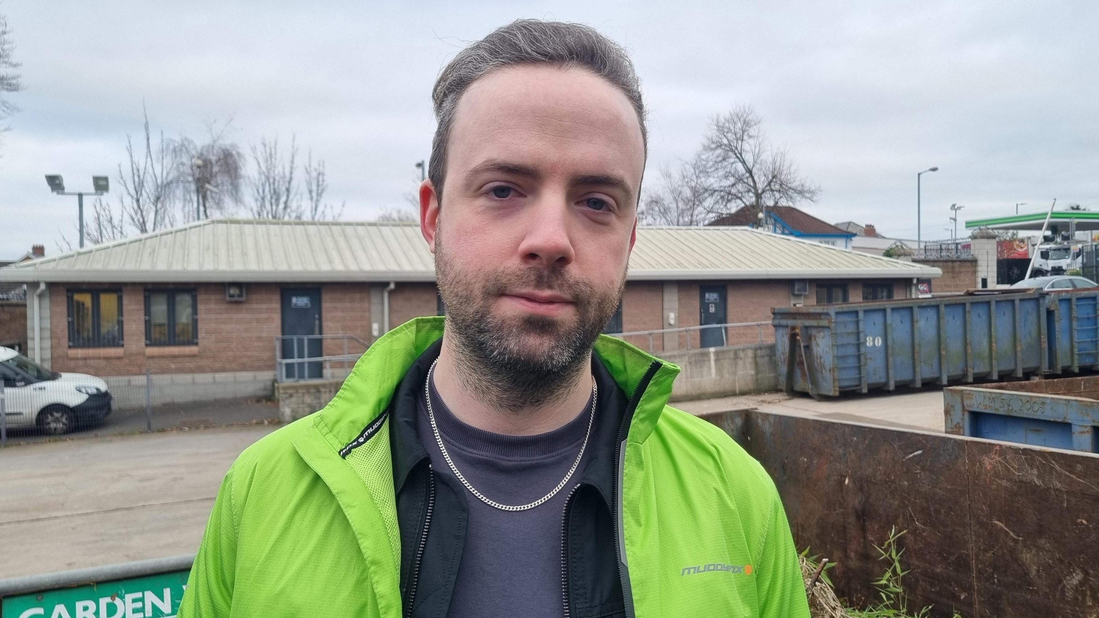 Green Party councillor Anthony Flynn at Palmerstown Road Recycling Centre in east Belfast. He has short grey-brown hair and beard and is wearing a bright green raincoat over a grey shirt and grey t-shirt. He is wearing a chain necklace