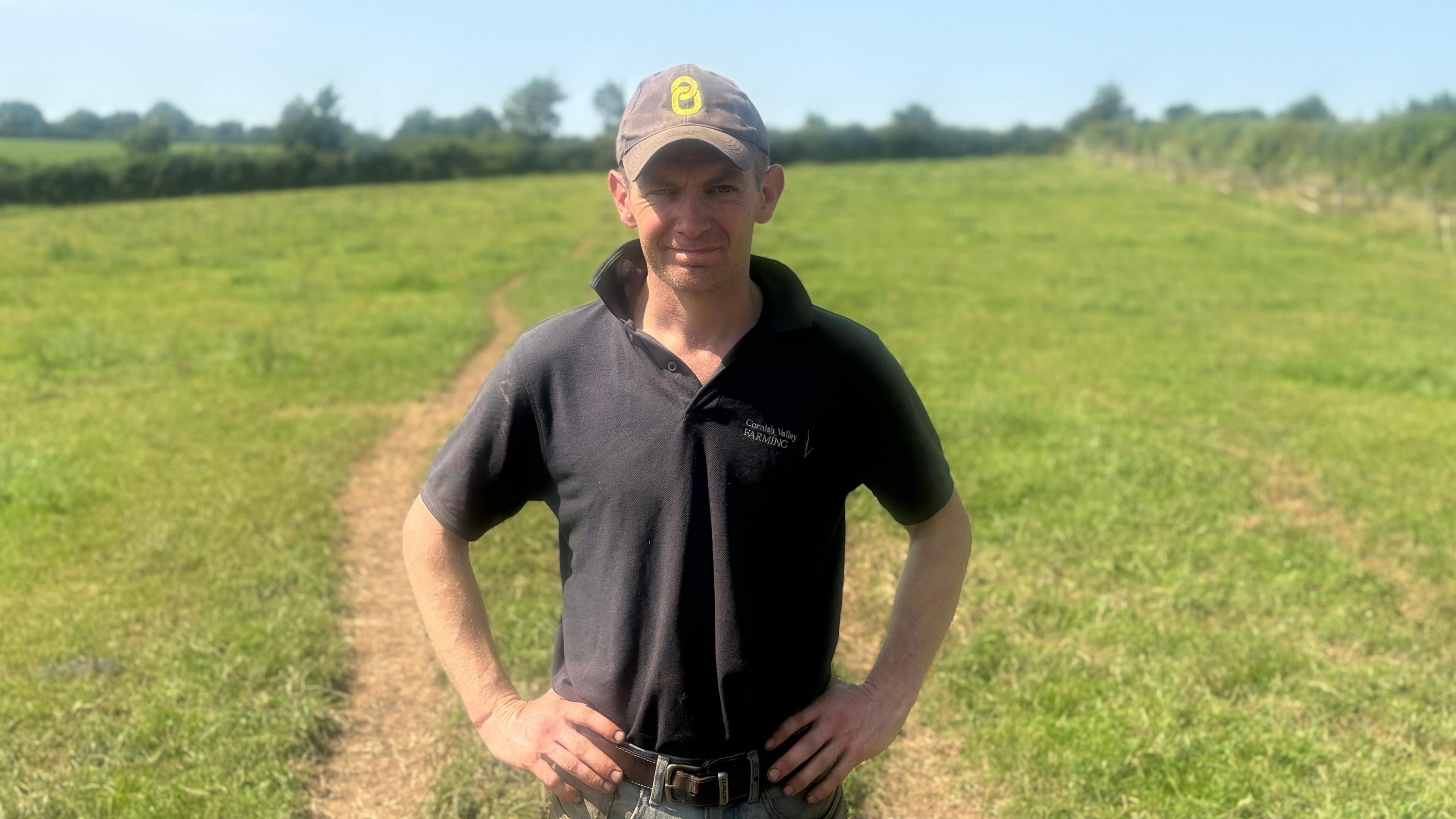 Rob wears a dark green polo shirt and a baseball cap and has his hands on his hips. He is standing in a grassy field with hedgerows and trees in the distance. 