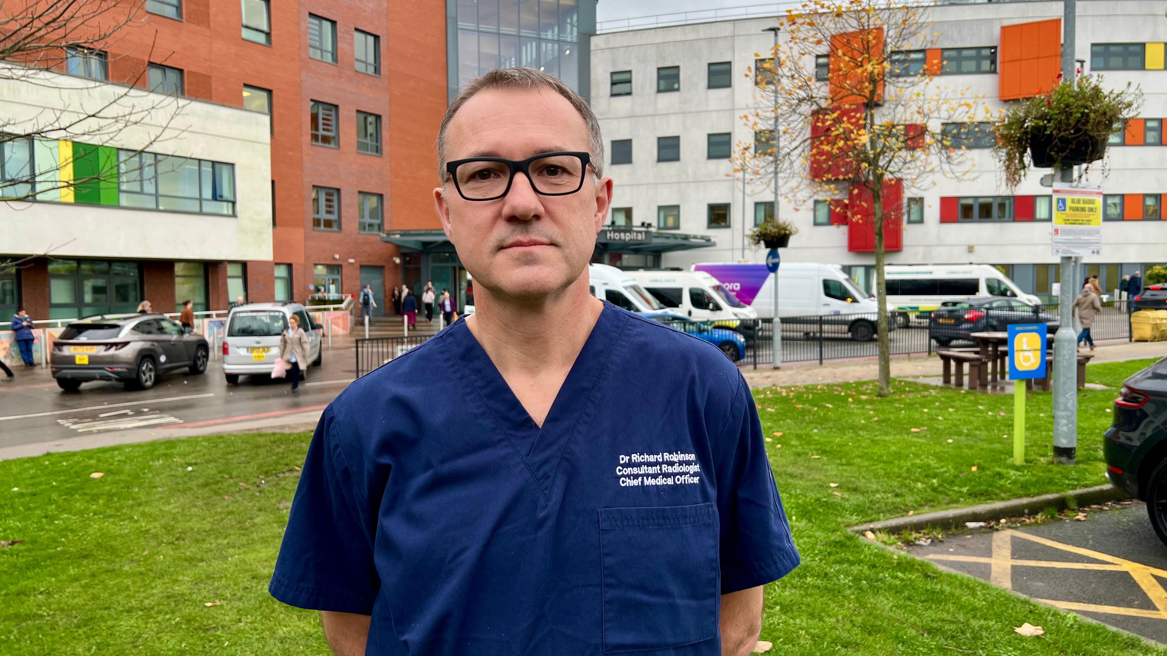Dr Richard Robinson is stood on grass in front of Pinderfields hospital in Wakefield. He is wearing dark glasses and dark blue scrubs