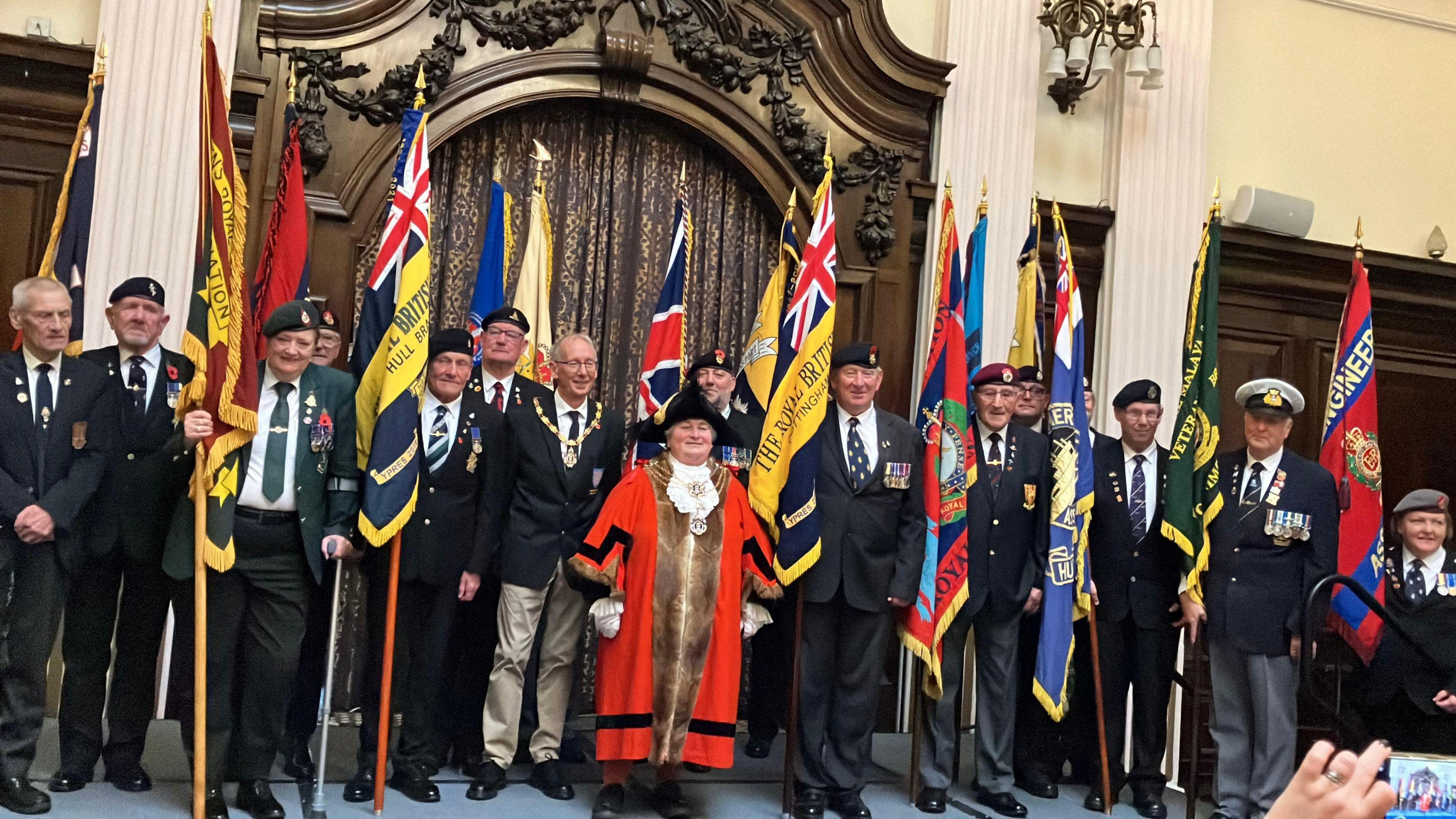 A group of 14 smartly dressed men and a woman, holding Royal British Legion flags and other standards, stand in a line in a grand hall. They wear regimental caps and ties and some have medals. In front of them is a woman dressed in red robes and a black admiral's hat.