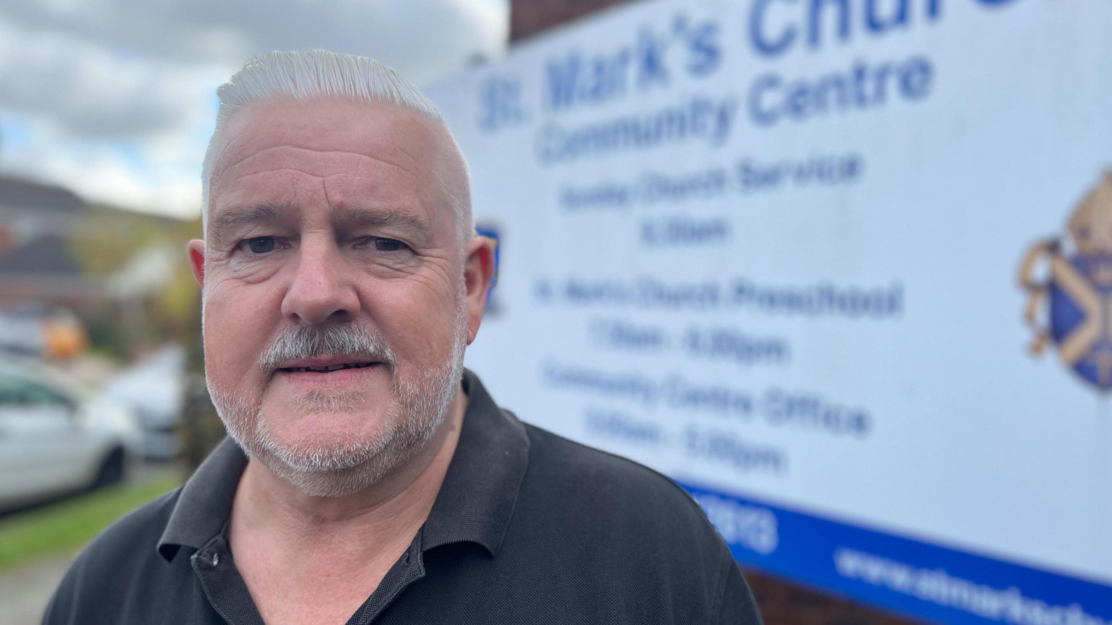 Charles Royden is wearing a black polo shirt and standing in front of a sign for St Mark's. He has short grey hair, stubble and moustache.