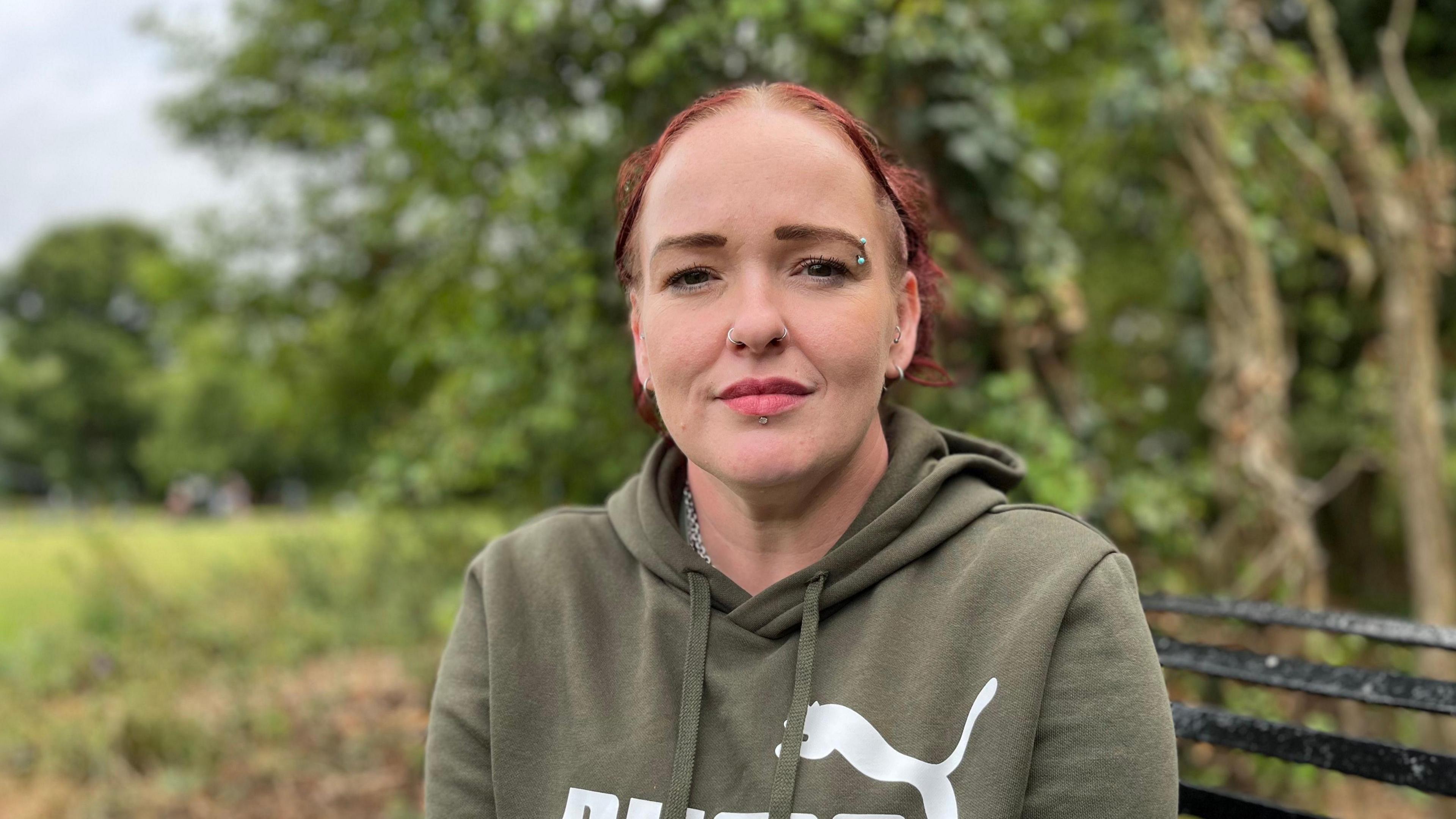 Rebecca Hinde is sat on a park bench, wearing a green jumper and has her hair tied back.