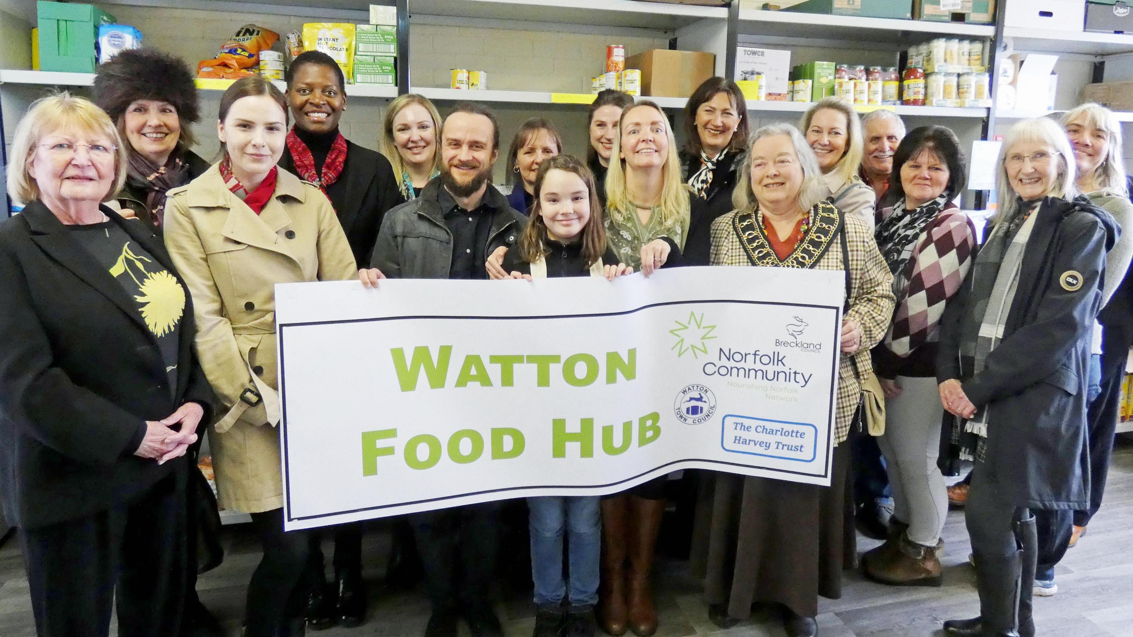 A group of people holding a banner reading "Watton food hub". There are roughly 15 people in the image. There are shelves of tins and other long-lasting food items behind them.