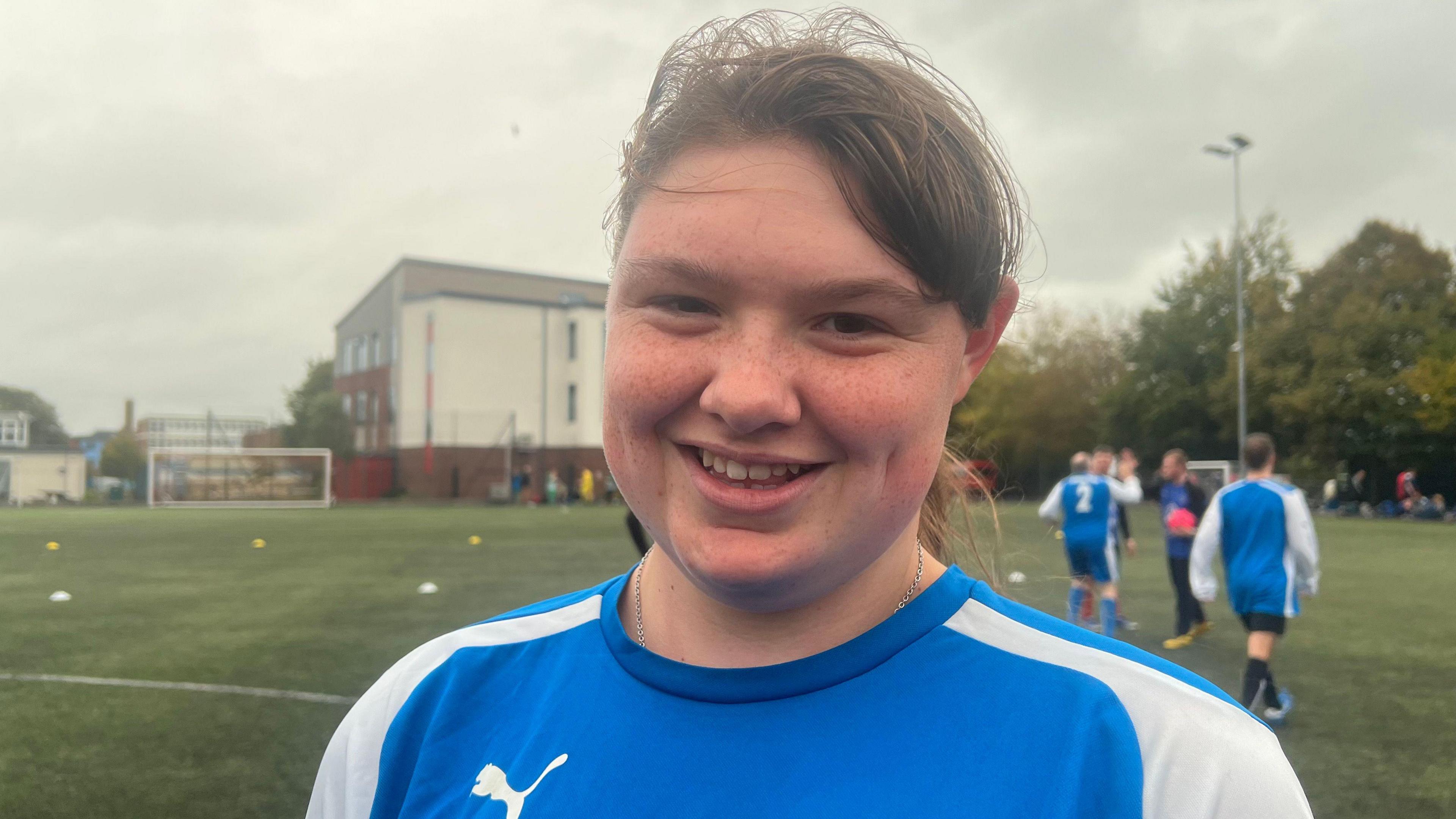 Emma in blue football kit standing on pitch with other players in the background.