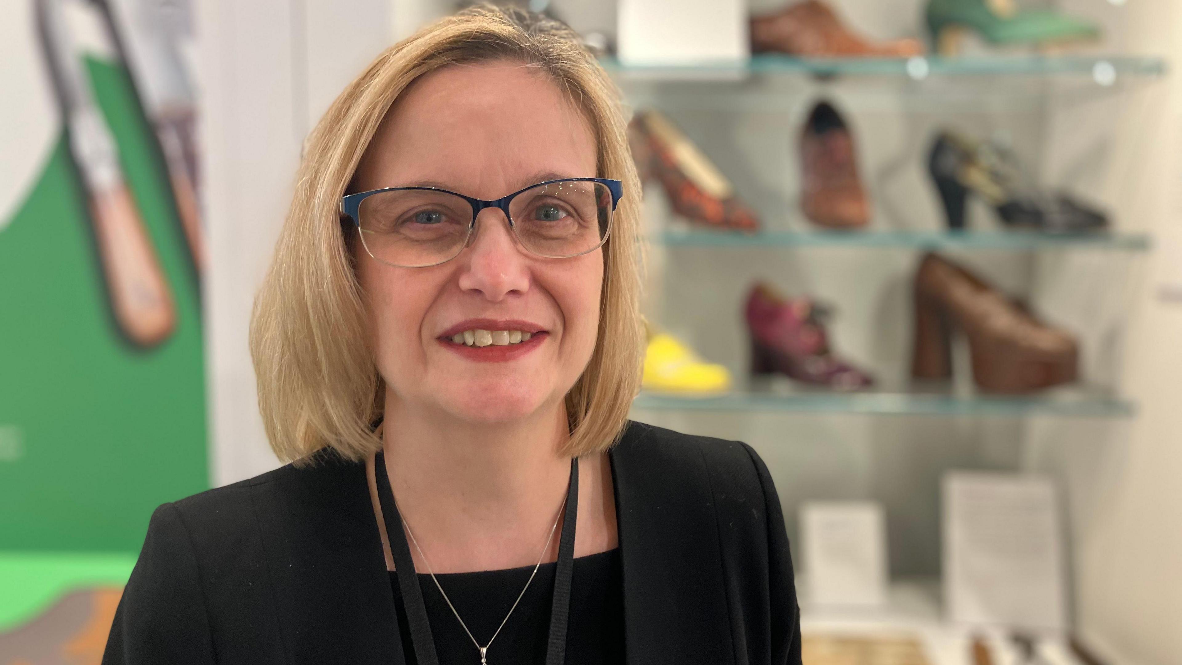 A woman with medium-length blonde hair and glasses, wearing a dark jacket, necklace, and black top, smiles as she stands in front of a display of old-fashioned shoes.