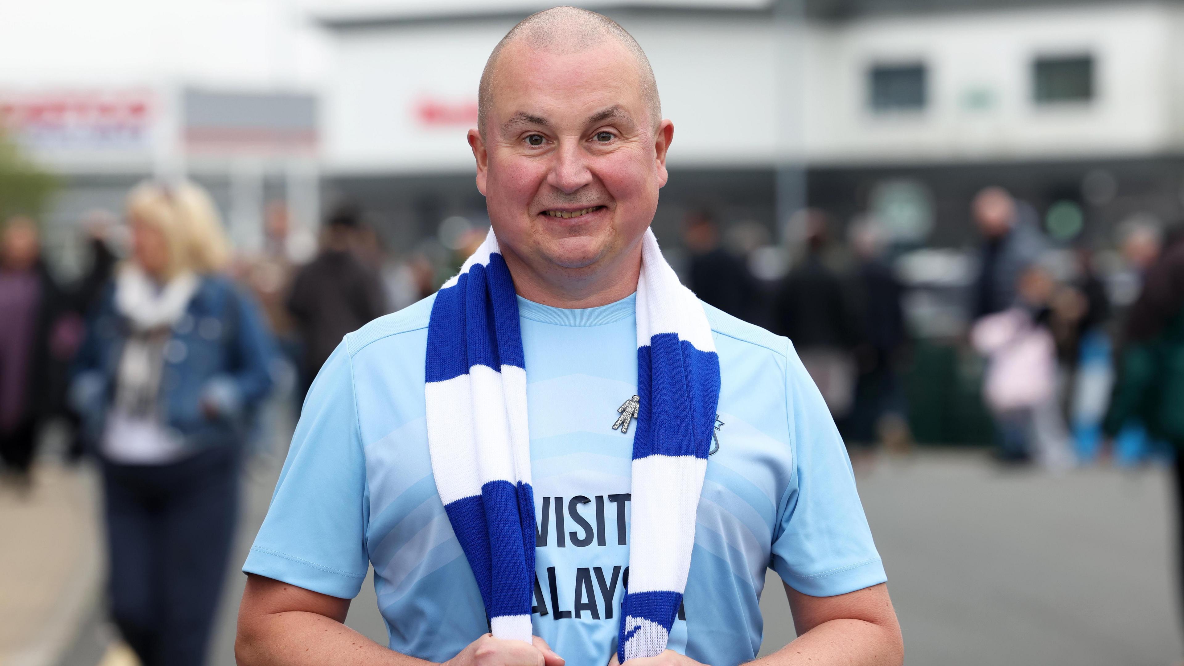 Mark Lewis is wearing a Cardiff City shirt and scarf, standing outside the stadium. He is smiling.