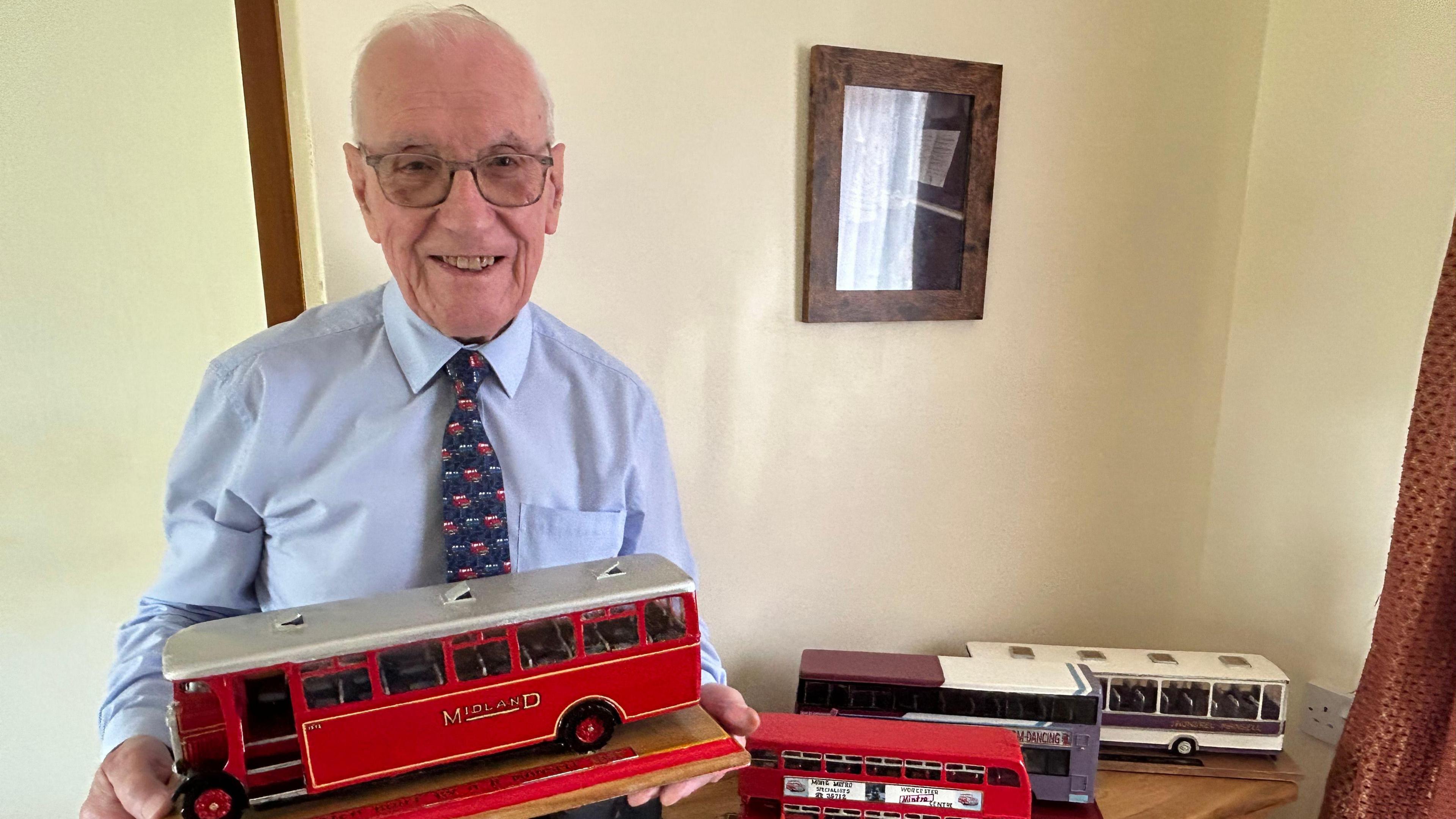 Arthur Mansell has white hair and glasses. He is wearing a blue shirt and blue and red tie. He is holding a red replica bus model. On a table next to him are three replica buses.