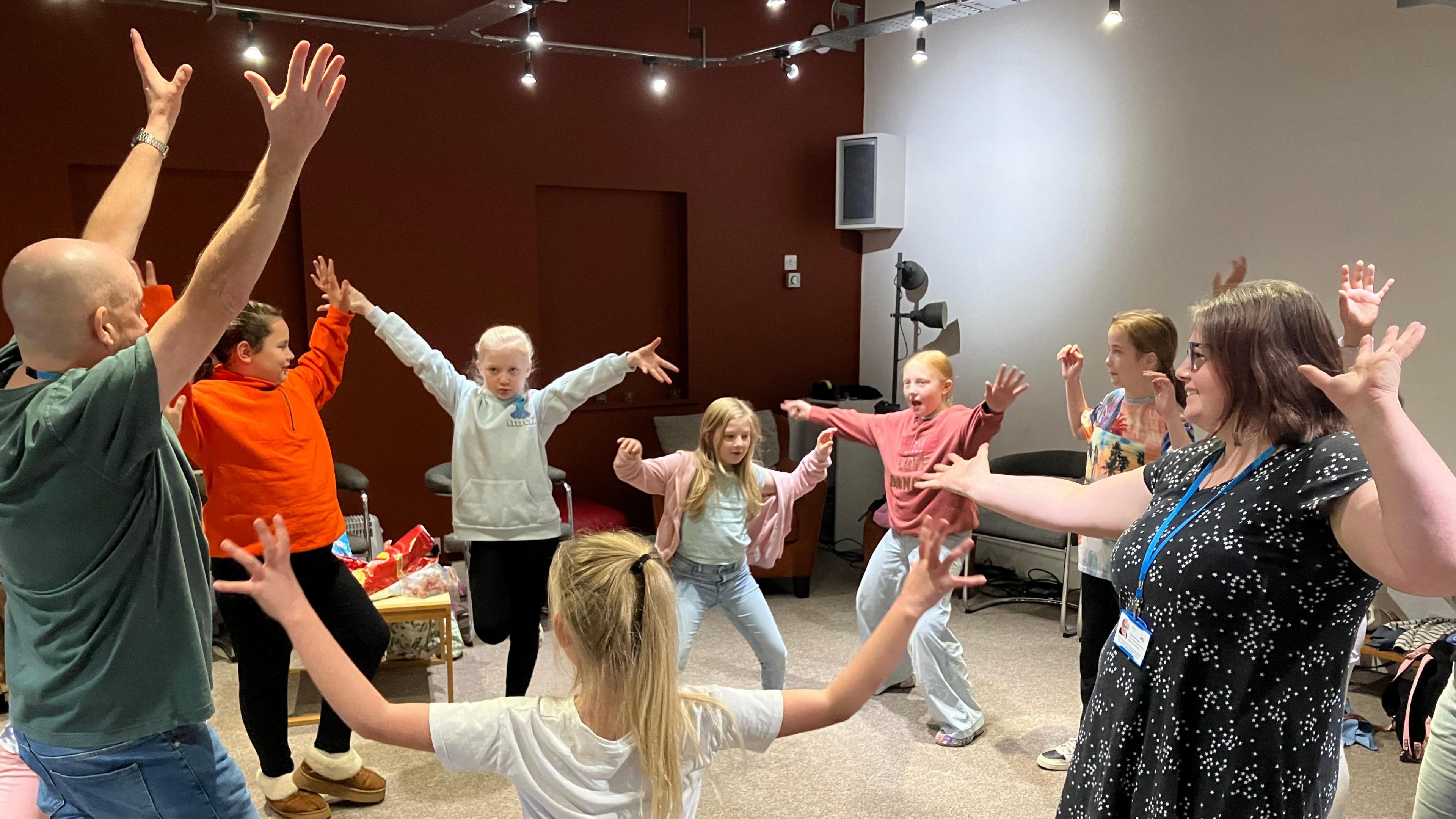 A group of children participating in a drama workshop by posing in different positions. They are all standing around a circle in a brightly lit room. Behind them are some chairs and snacks on a table. 