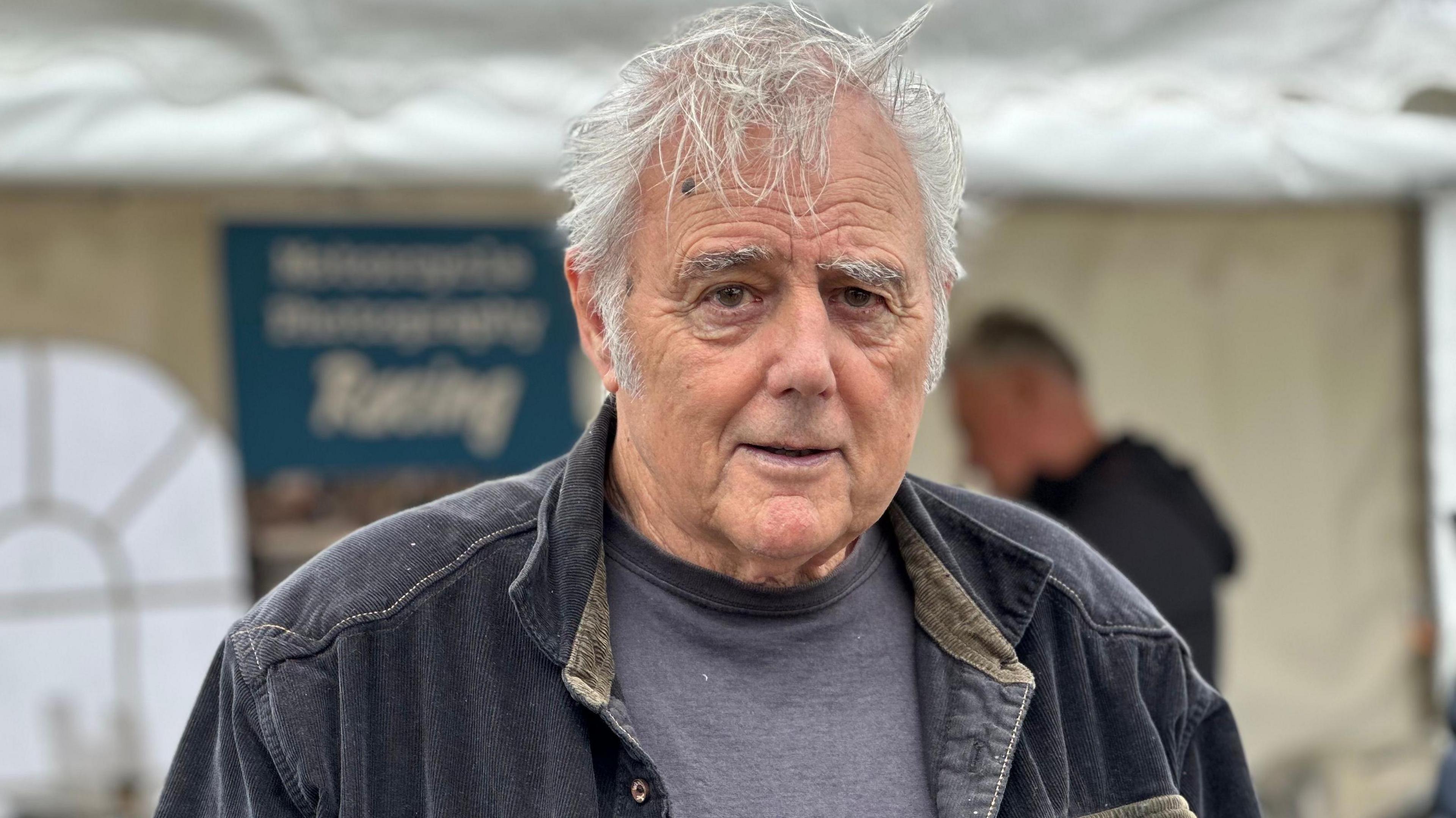 Dave Lewis stands in front of a white marquee, he wears a navy corduroy jacket and grey T-shirt. He has grey hair that is blowing in the wind.