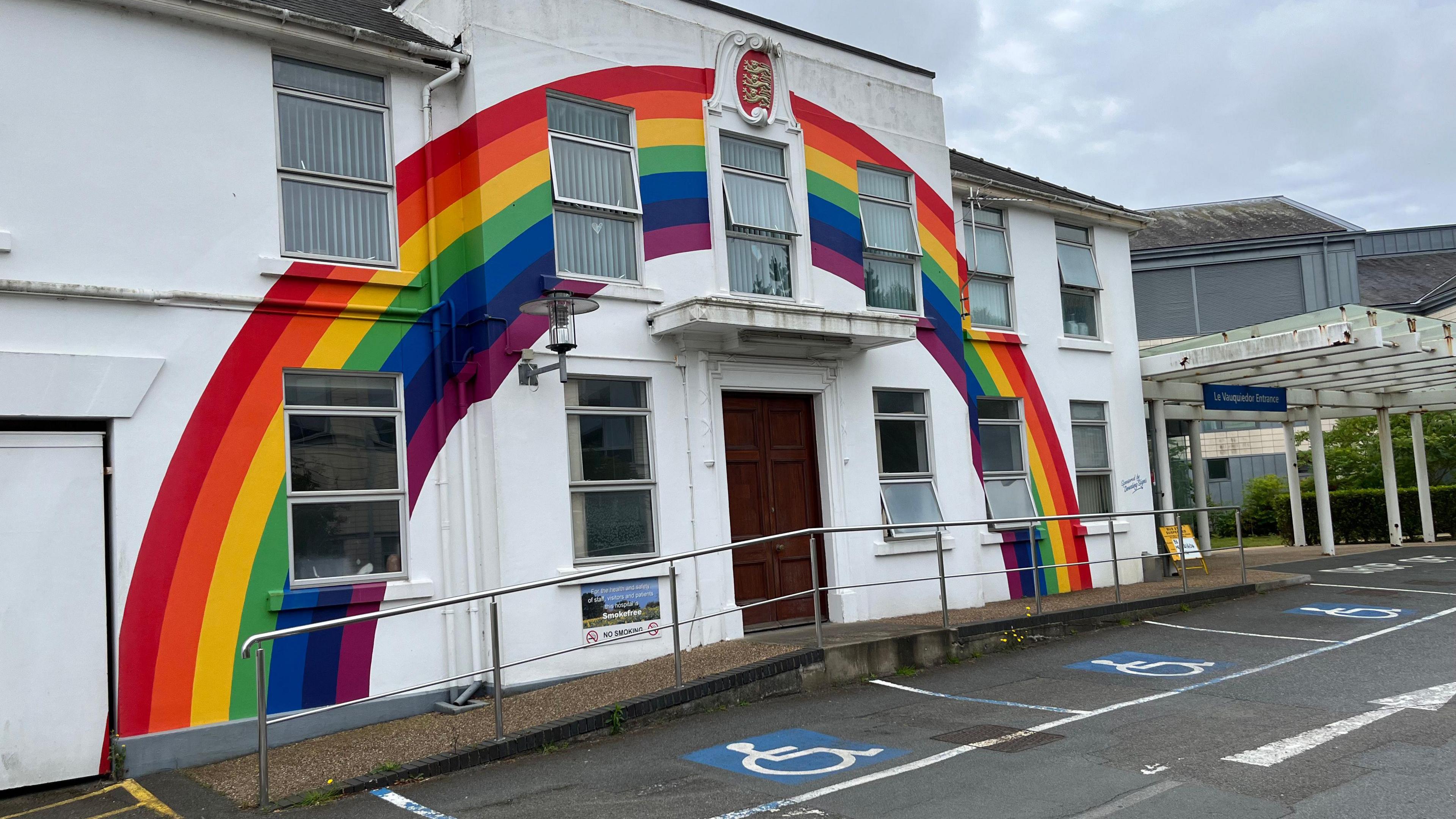 A white building with a ranbow painted on it. There are three accessible parking spaces in front of the building. 