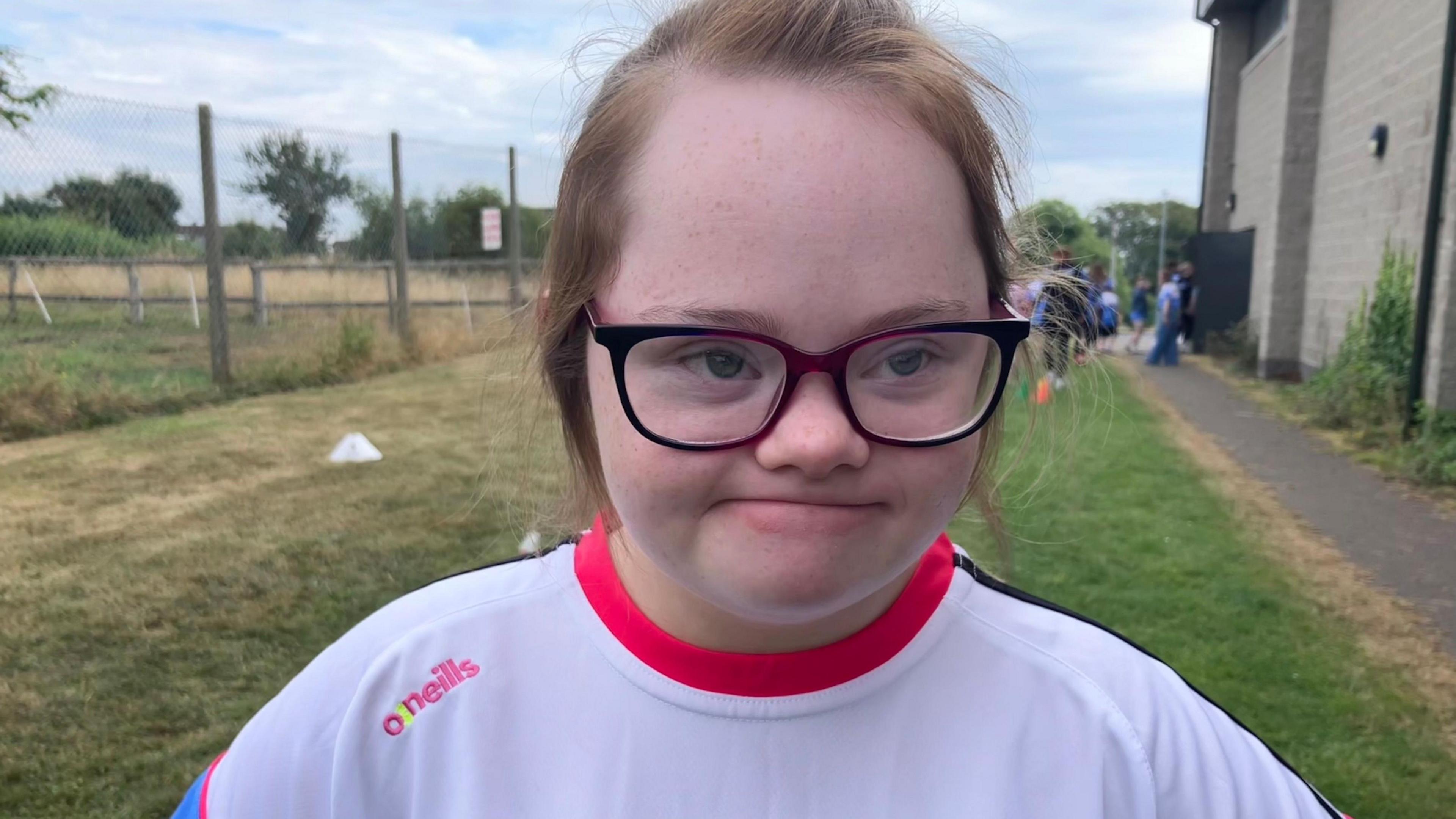 Leah has fair blonde/ginger hair. She wears glasses and a white top with a pink collar and black stripes across the shoulders. Halfway down her upper arm here clear turn blue. 
