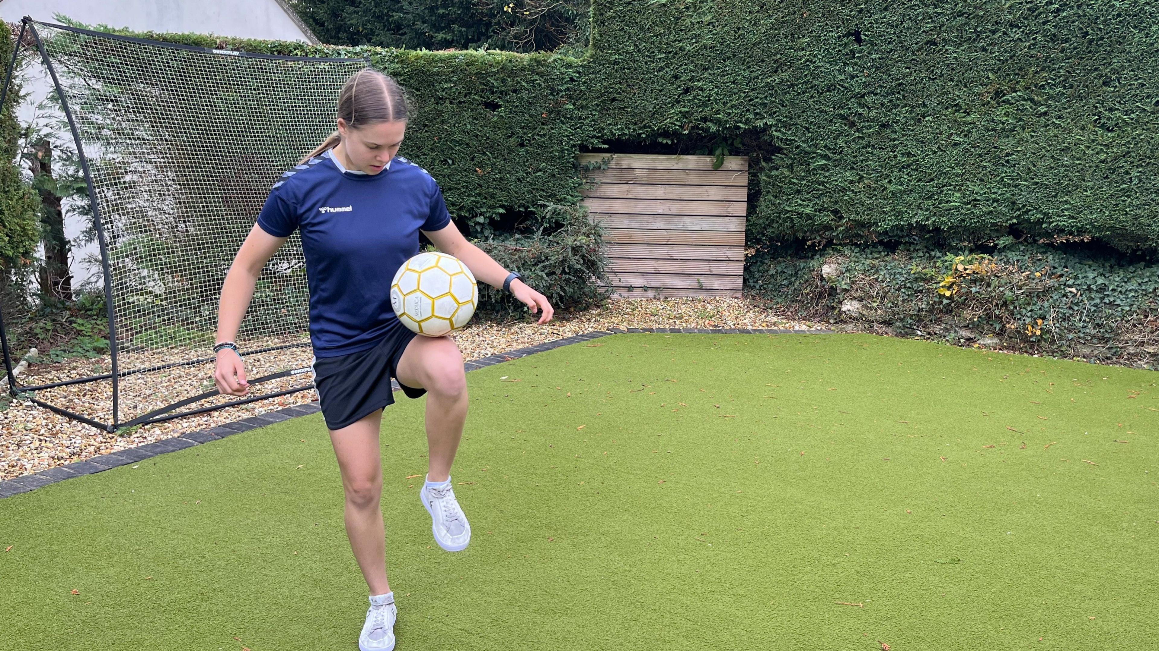 Isabel Wilkins in the middle of a freestyle football trick. She is wearing white trainers, black shorts and a dark blue football jersey. She is balancing the football on her left knee while standing on the other leg, with her arms out beside her. She is in a back garden with grass, neatly cut hedges, a wooden fence and a football net.