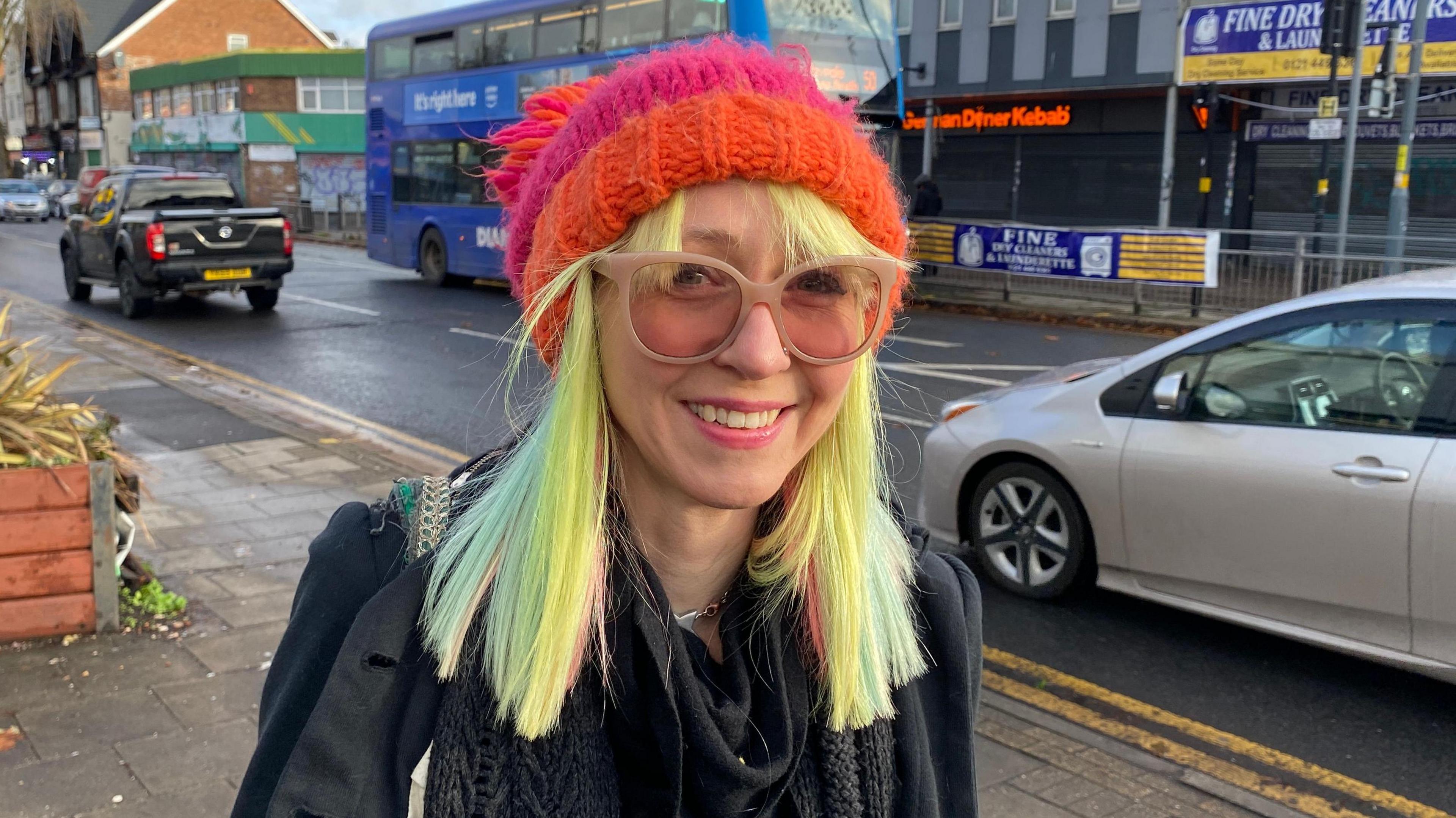 Victoria Platt pictured smiling weating a pink and orange hat and a pair of brown glasses. She has yellow,green and pink hair that is shoulder length and wears a black jacket.