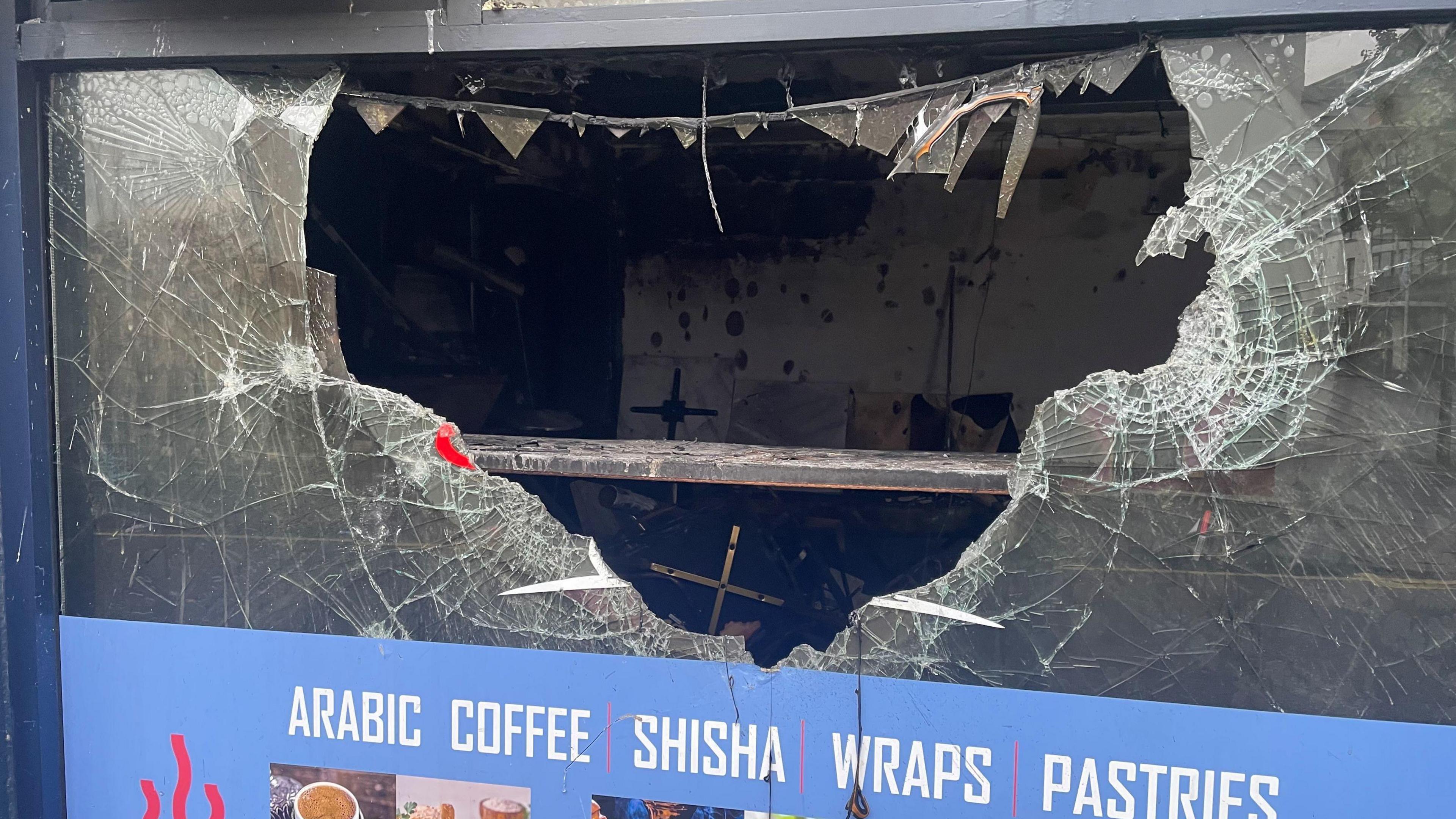 Cafe with a sign reading "Arabic Coffe, Shisha, Wraps, Pastries" the cafe's window is smashed