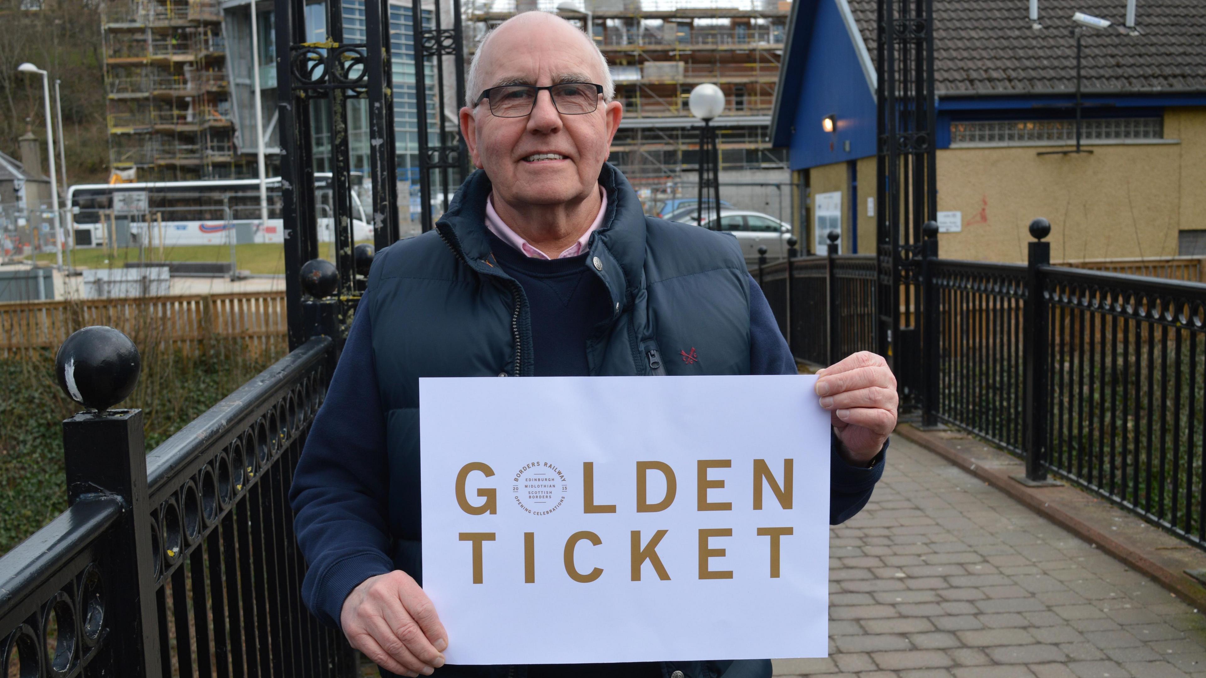 A bald headed man in glasses in a blue jacket and jumper with pink shirt underneath holds up a piece of paper with the words "golden ticket" on it
