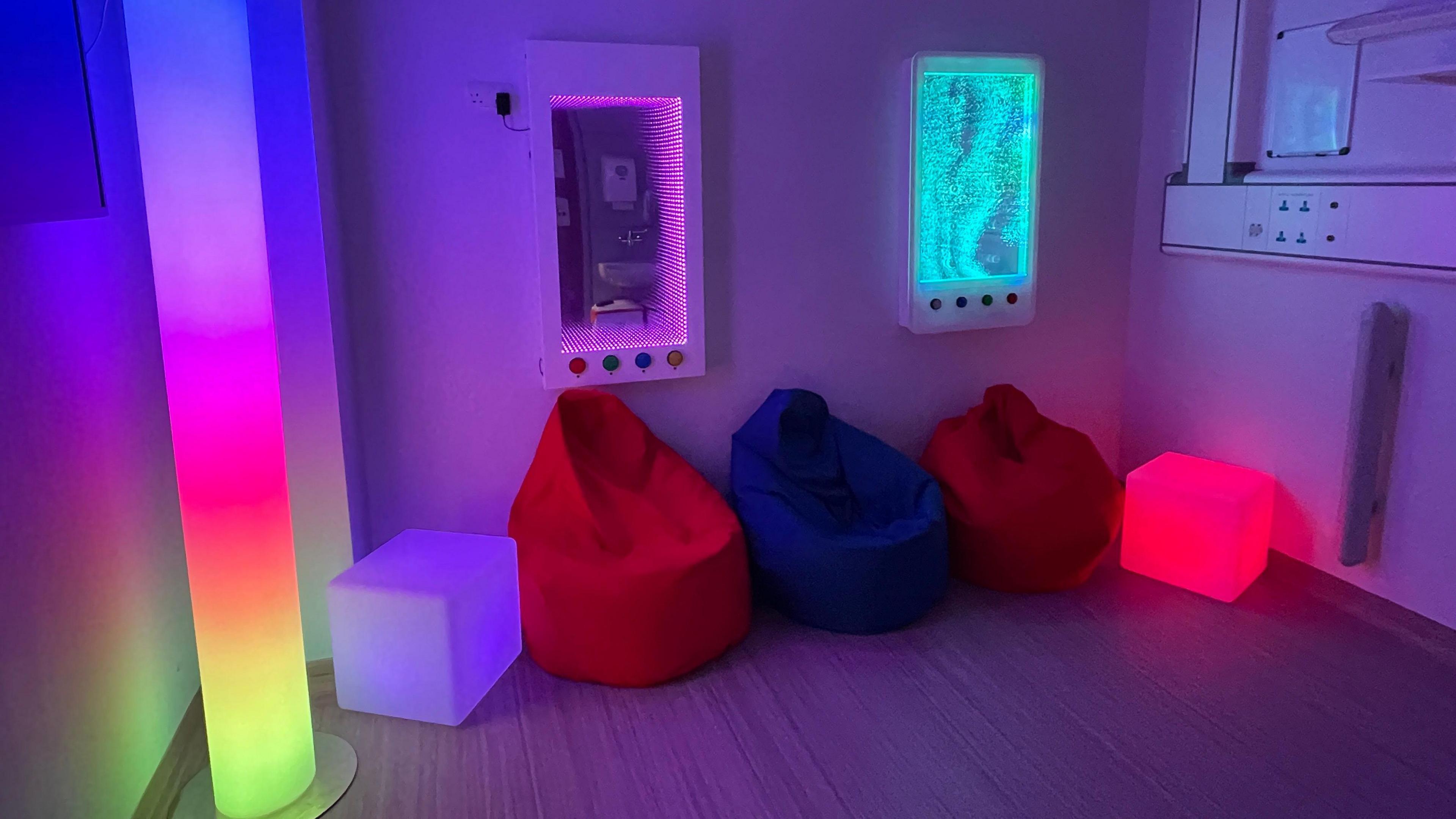 Three bean bags sit in a room, two boxes sit either side and a pole with colourful lights sits on the far left hand side. A white wall also features colourful lights. 