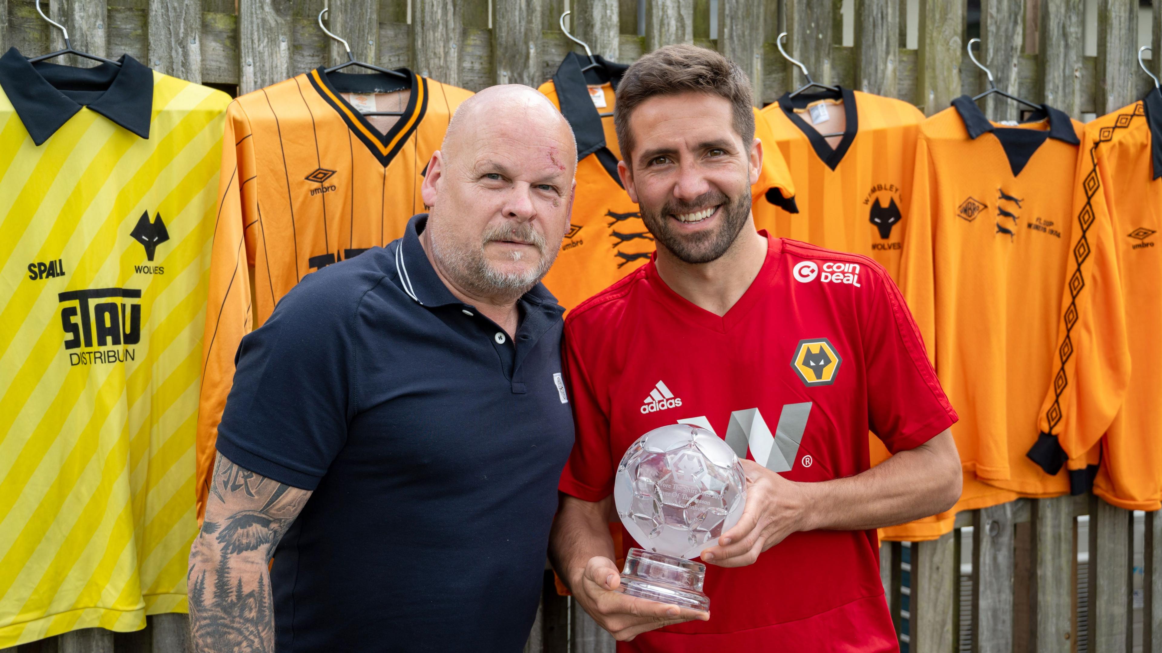 One man in a blue polo shirt, one man in a red football shirt holding a sporting trophy with six football shirts hanging in the background