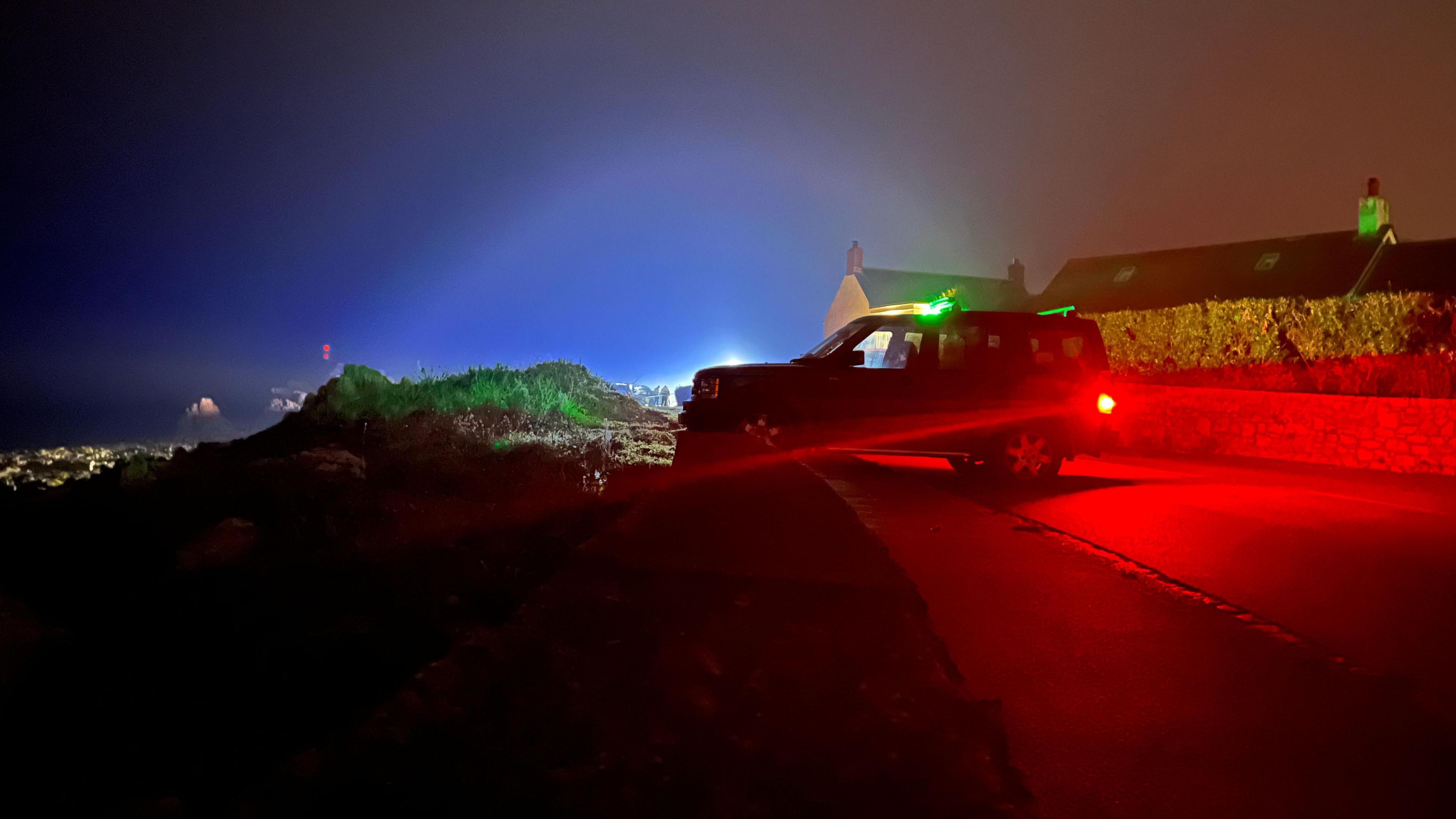 A vehicle is parked across the road at night. Behind bright lights can be seen.