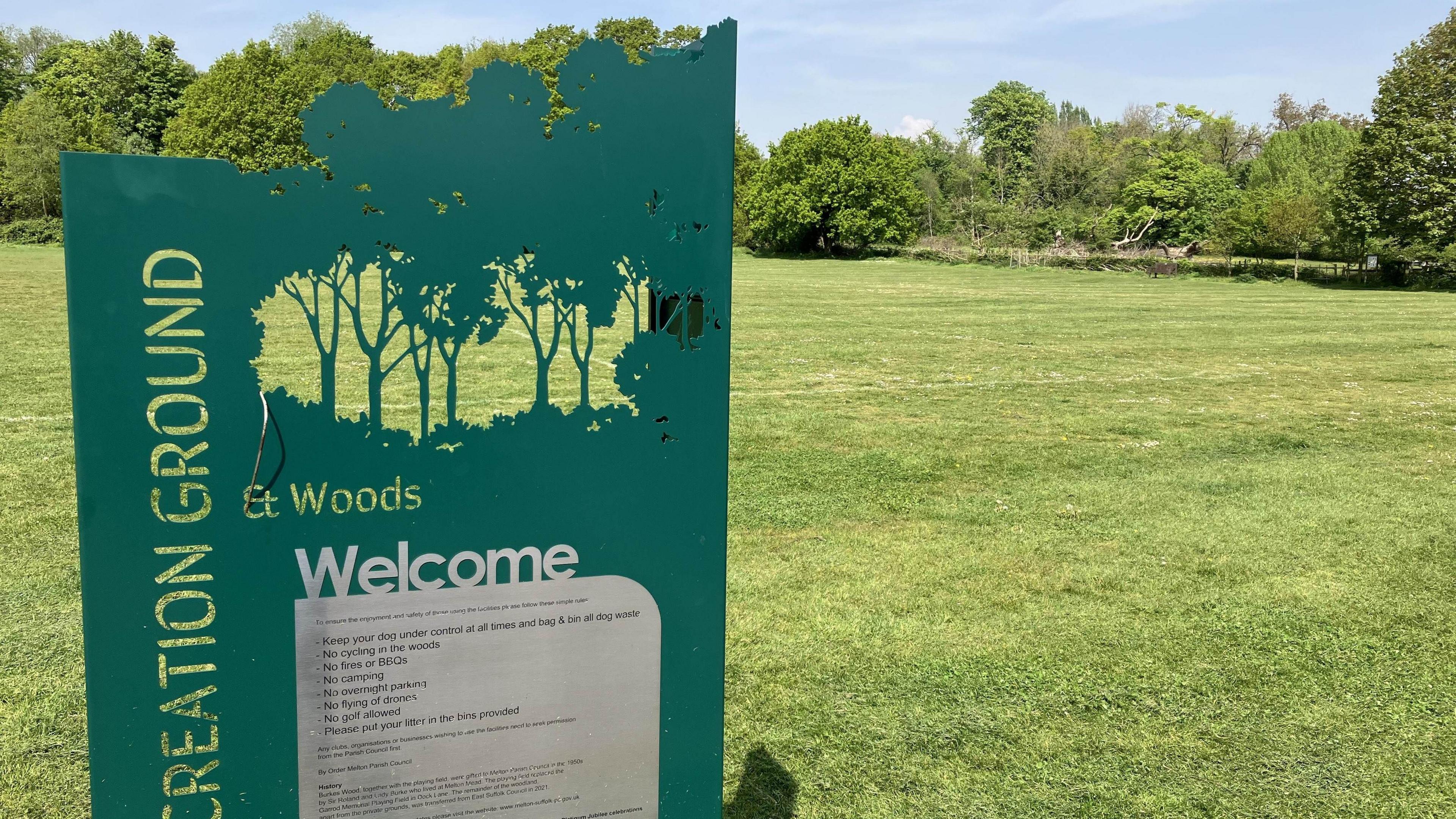 Green sign post at Melton Recreation Ground and Woods in front of large grass area  