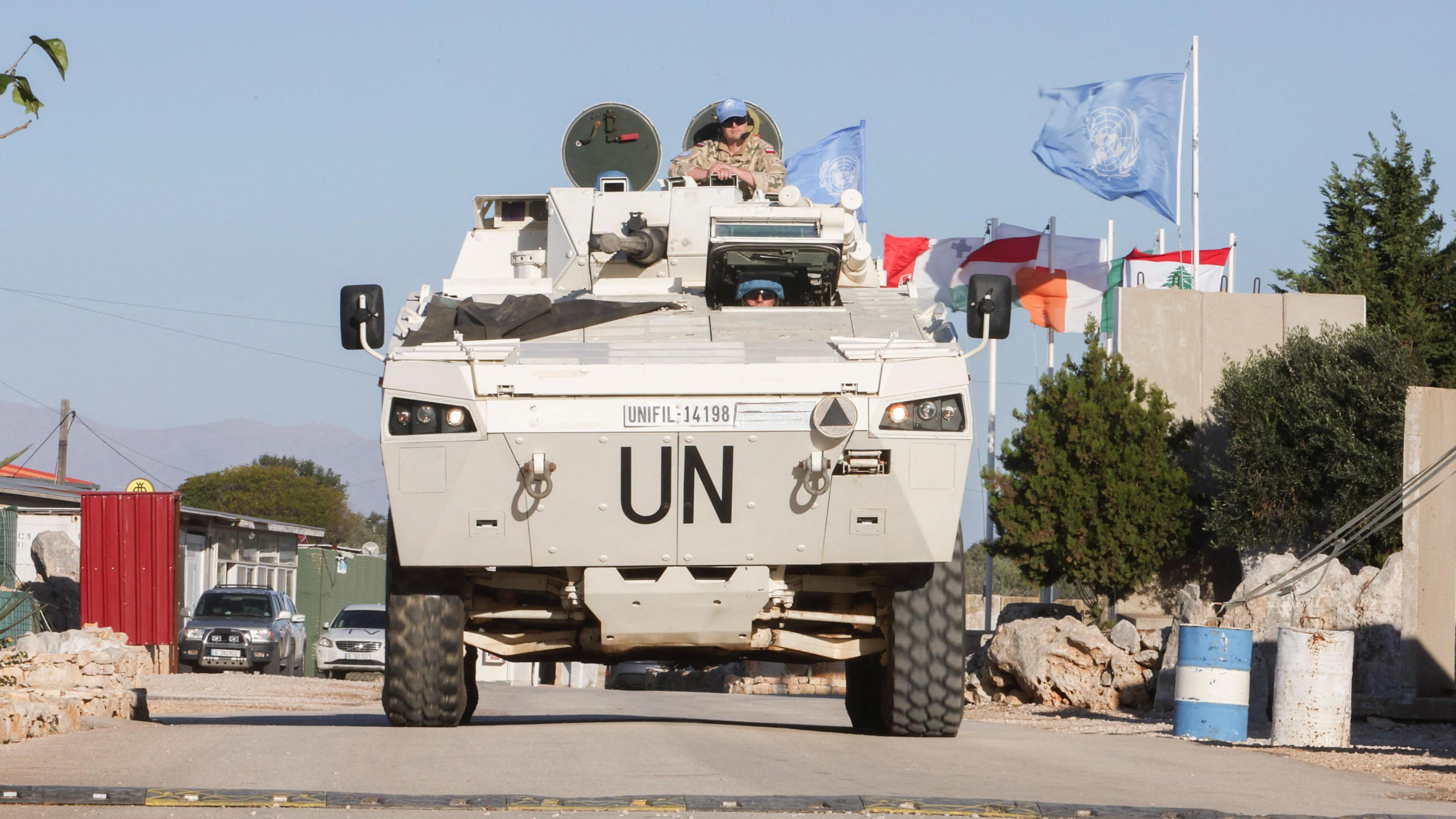 Army tank driving through camp shamrock in southern Lebanon, UN peacekeeper pictured on top of tank, several countries flags in background