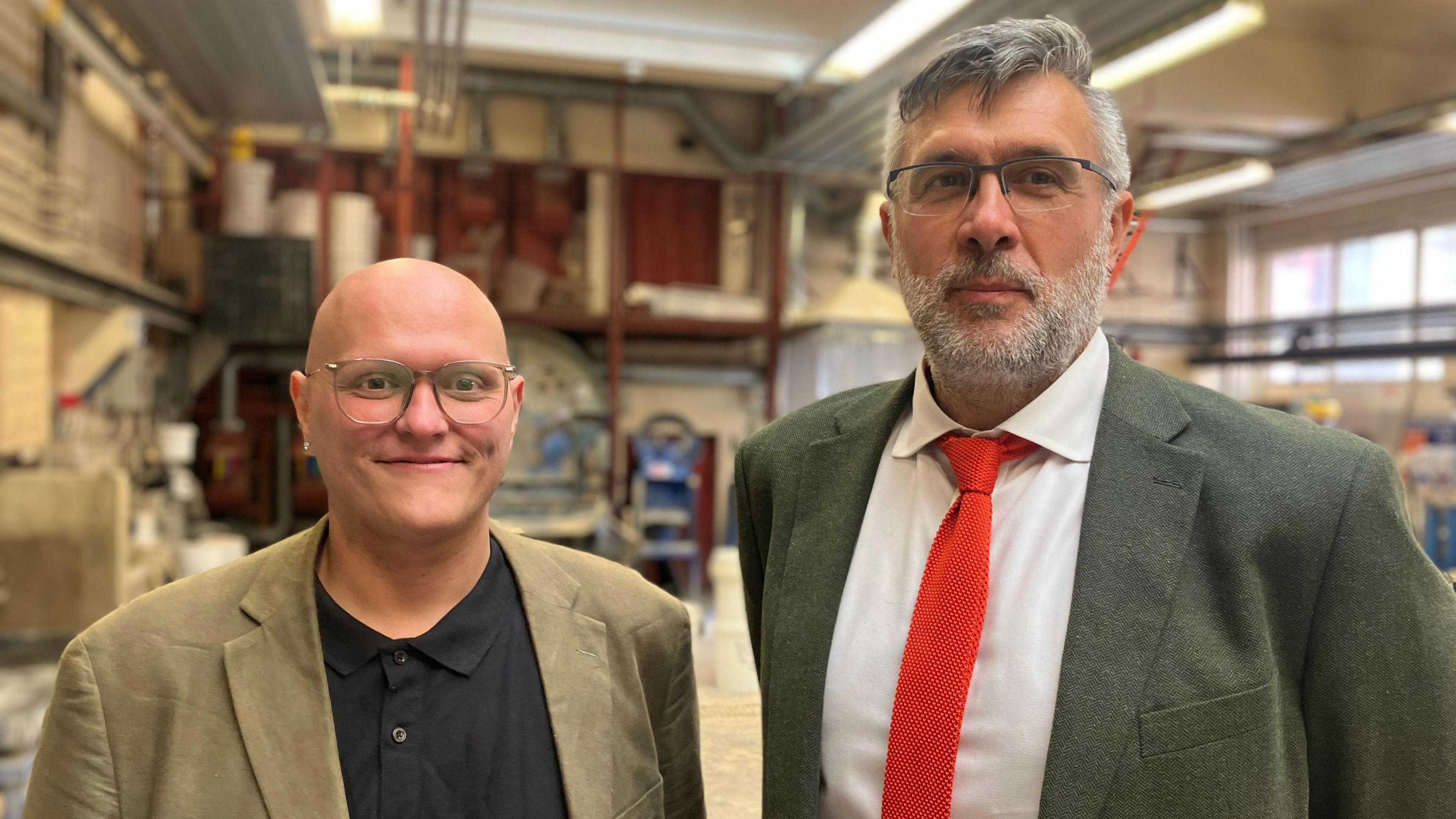 Two men smile at the camera. One man wears a black shirt with a green blazer. The other has a grey beard and wears a white shirt, orange tie and green blazer.