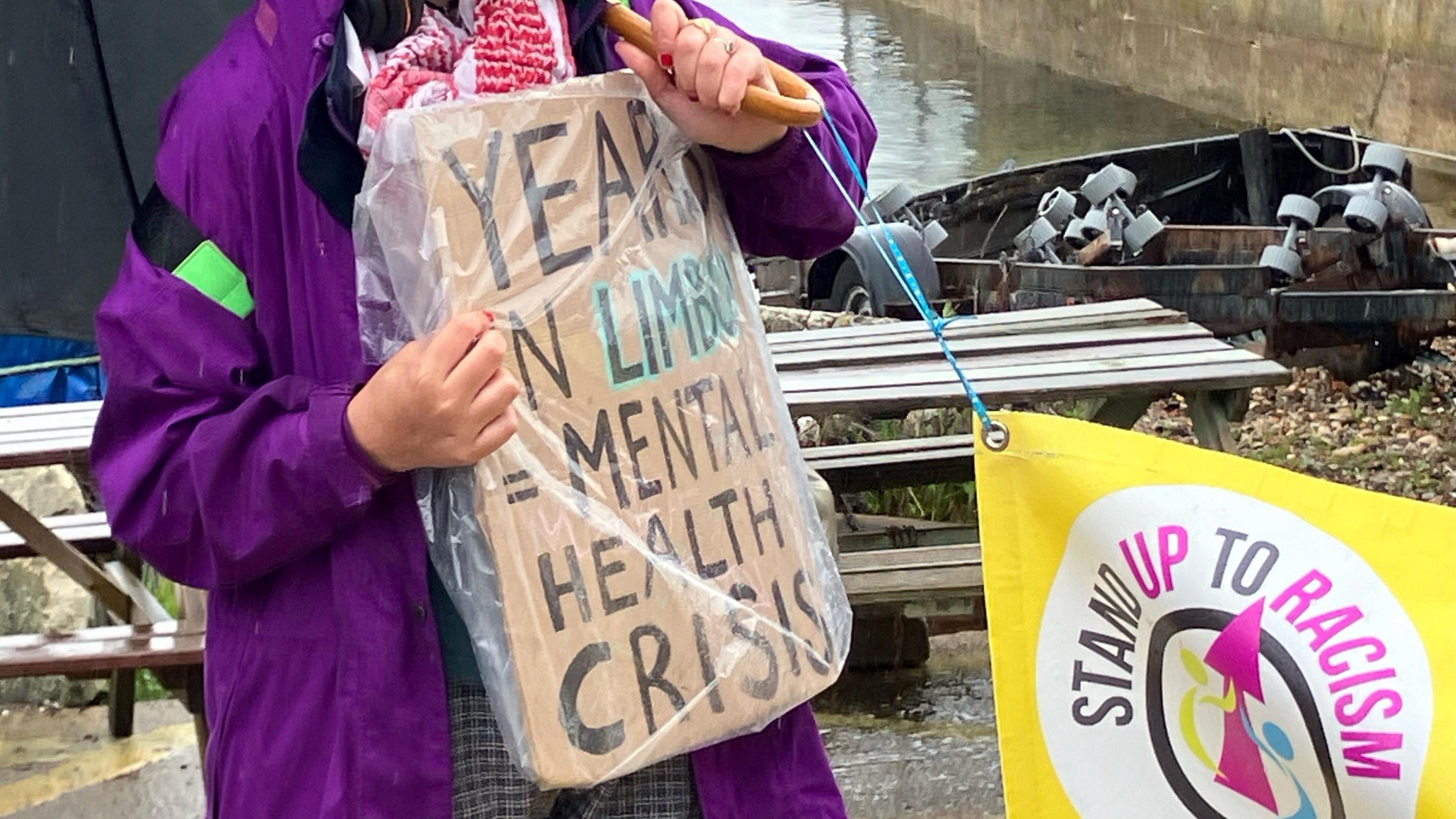 A woman wearing a purple coat holds a placard that reads: "Years in limbo = mental health crisis"