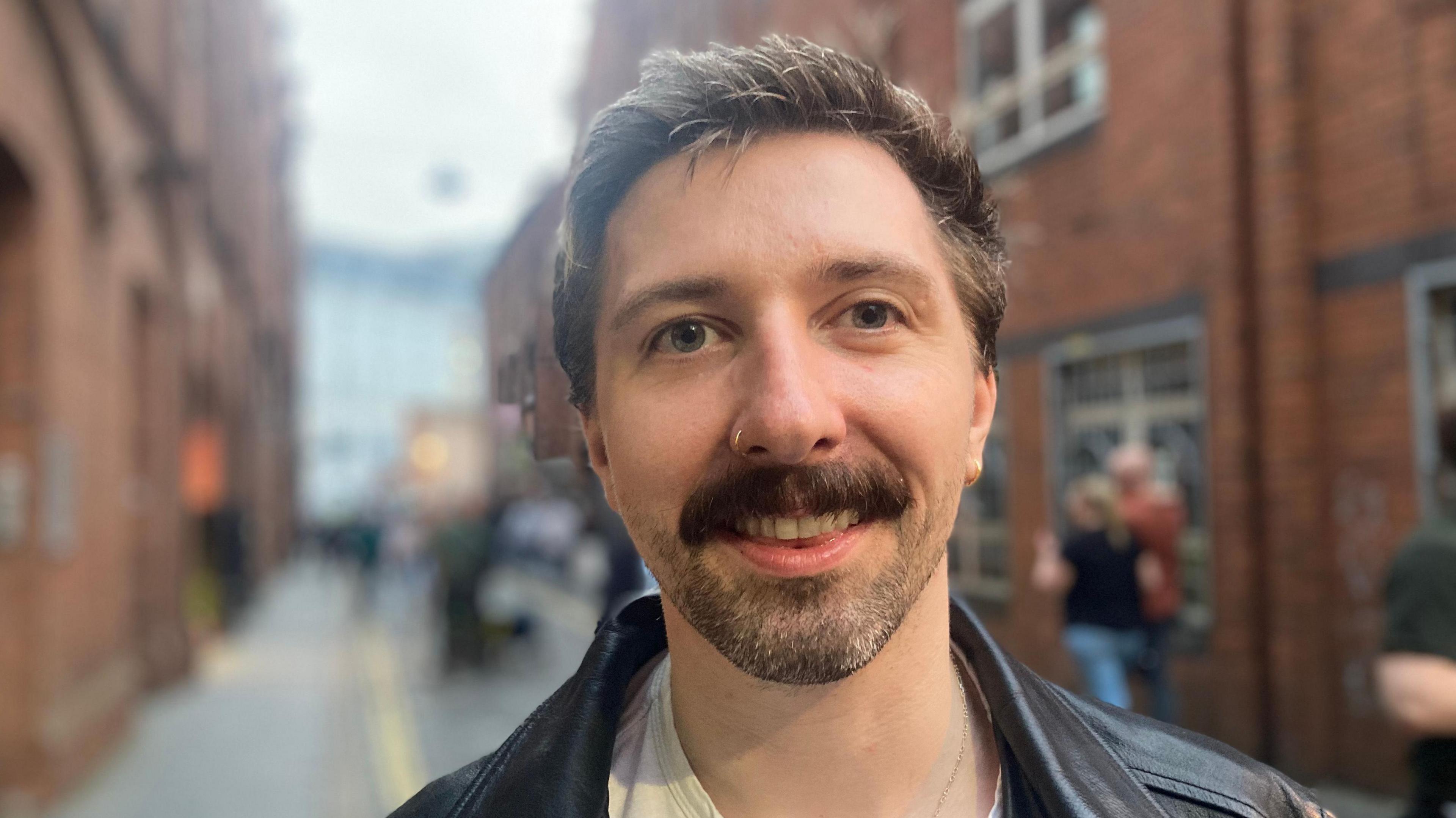 Pete, a man with short darkish hair, a moustache and stubble, looking into the camera on a narrow Belfast street lined with red bricked buildings.  He is wearing a black leather jacket. 