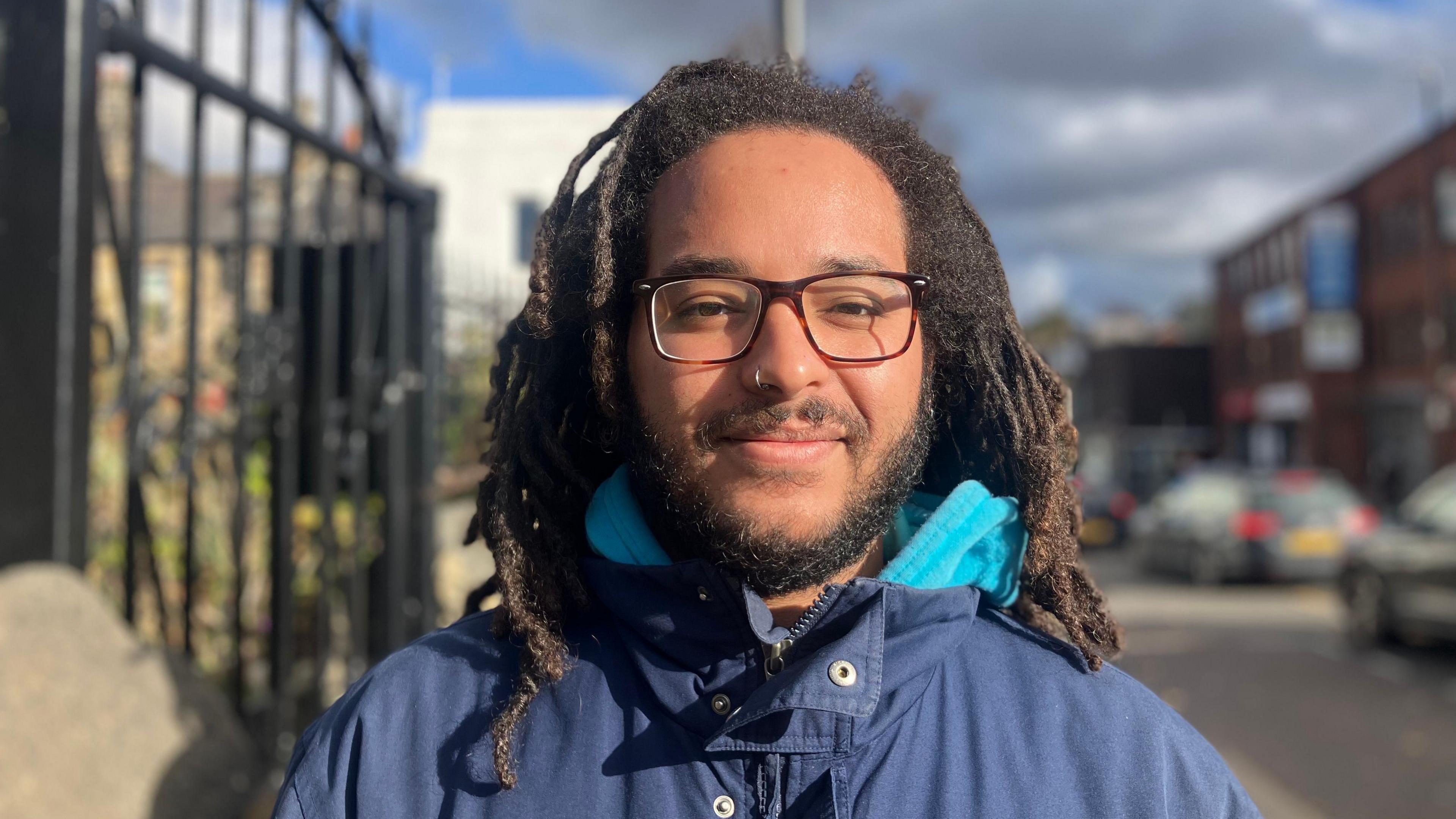 A man with dreadlocks, a beard and glasses smiles. He wears a blue coat. Headingley is blurred behind him.
