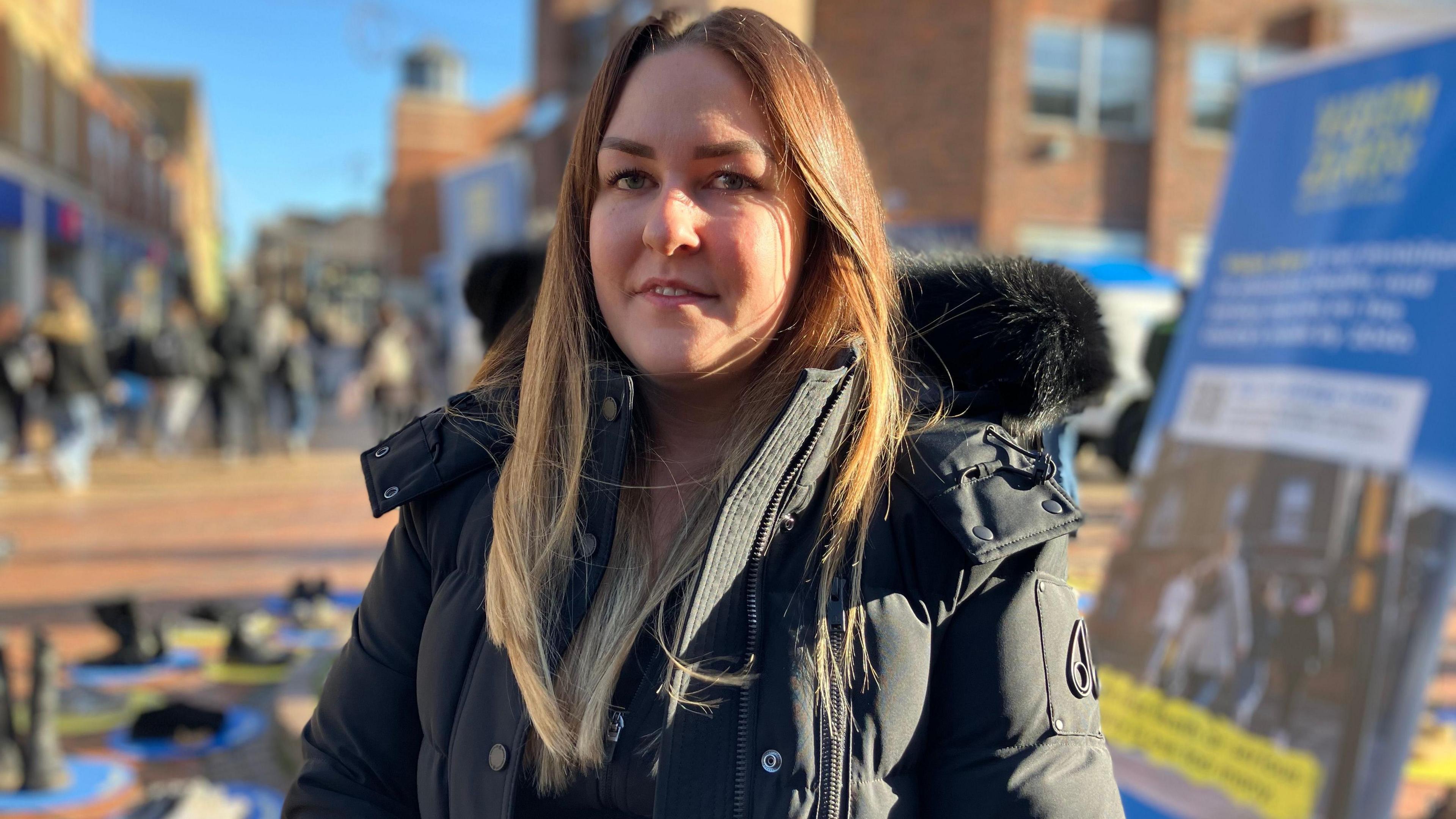 A woman in a black coat with a busy high street in a soft focus behind her.