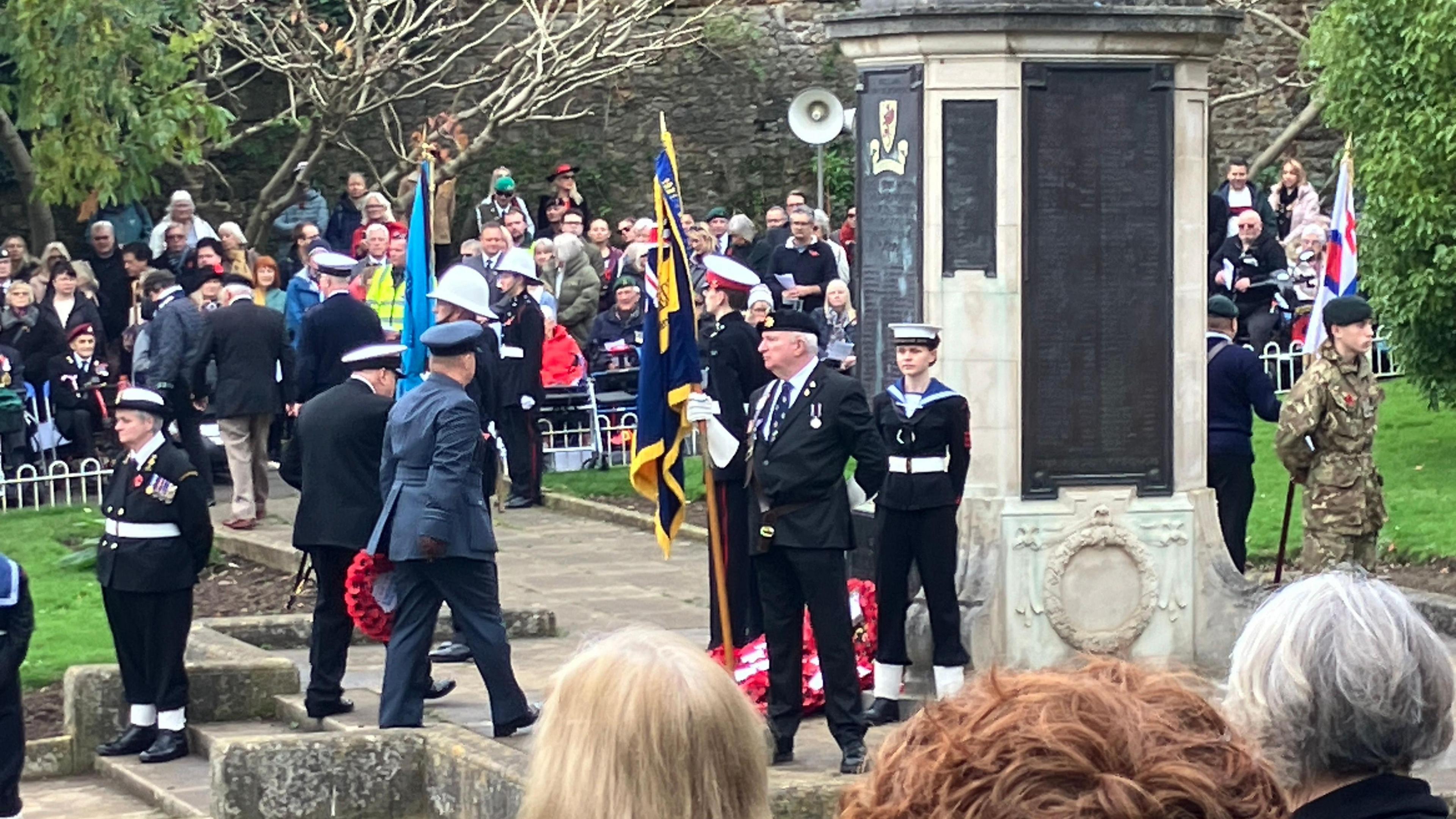 Military officials approach the war memorial in Grove Park, with hundreds of people watching on. There are flag bearers in uniform standing around the memorial and wreaths of red poppies have been laid at its base. 